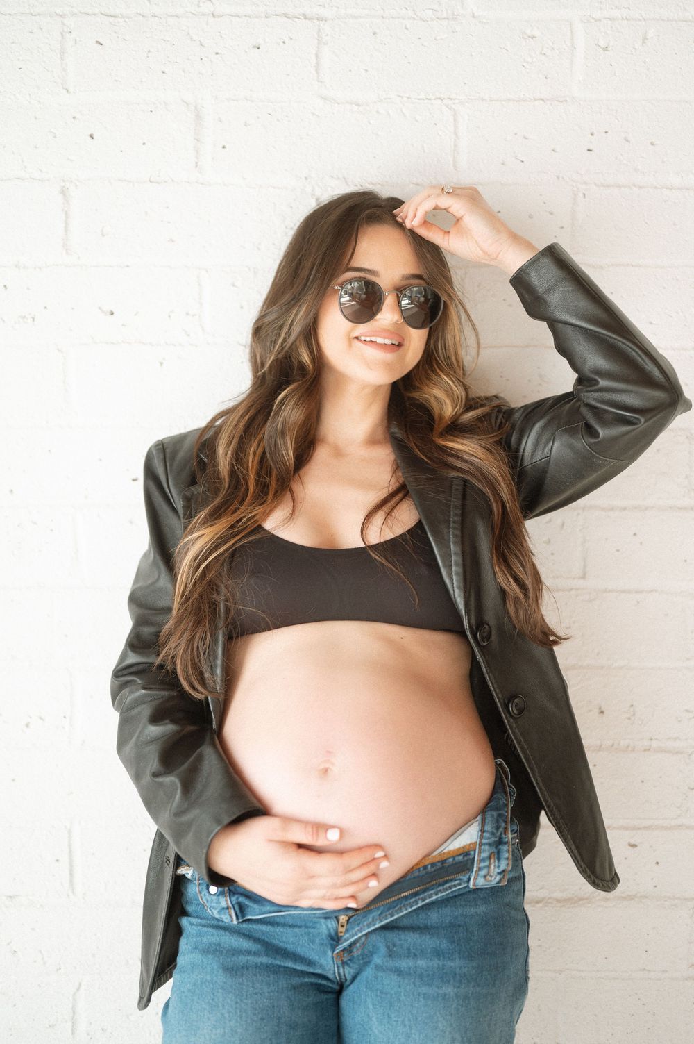 A series of maternity photos showing a casual look with a leather jacket, black top and jeans against a white wall.