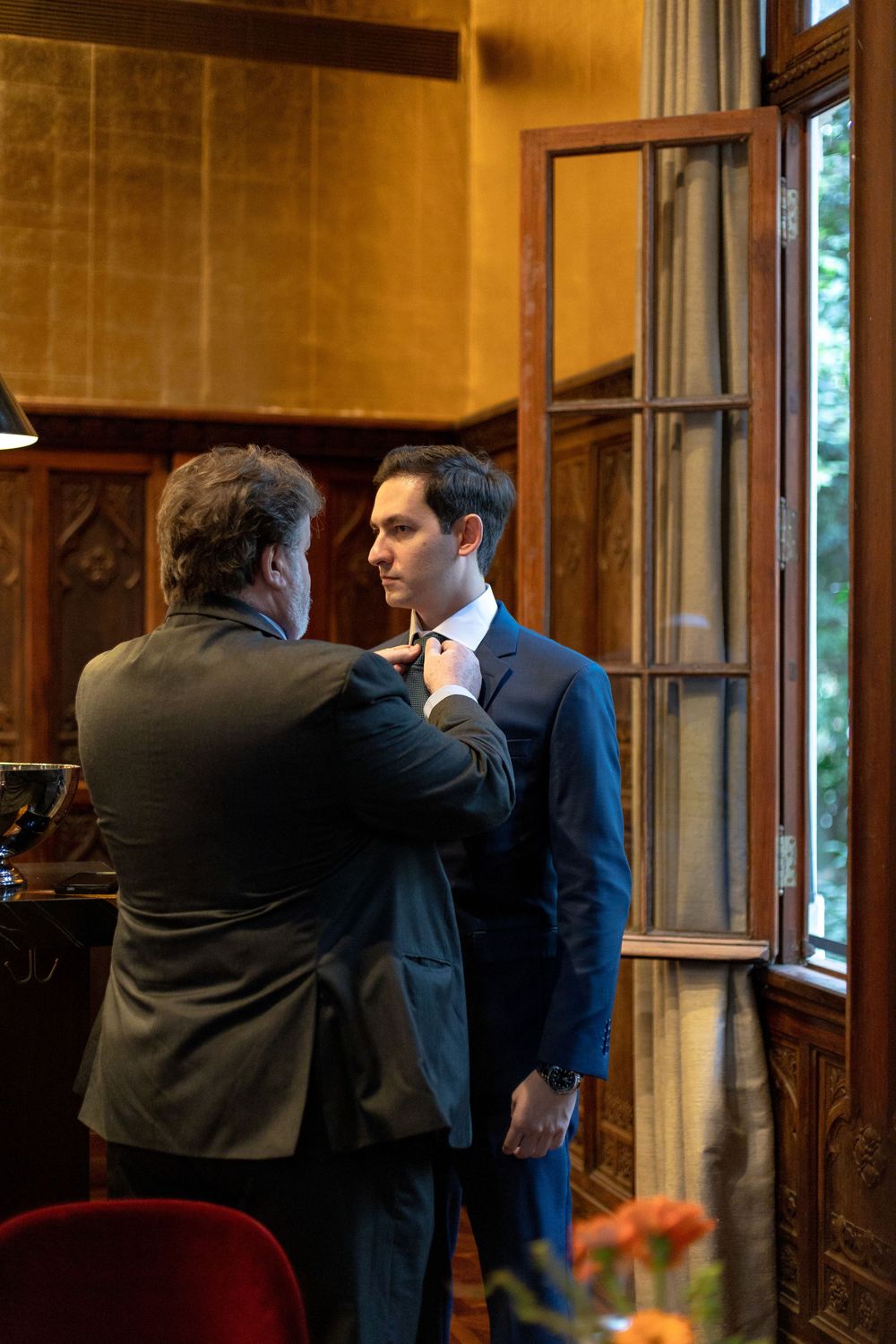 Two people in business attire having a conversation in an elegant wood-paneled room with tall windows.