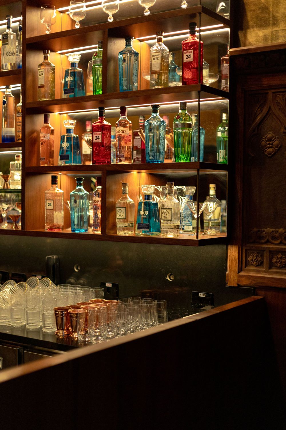 Illuminated bar shelving displays colorful liquor bottles and glassware in a dimly lit upscale establishment.