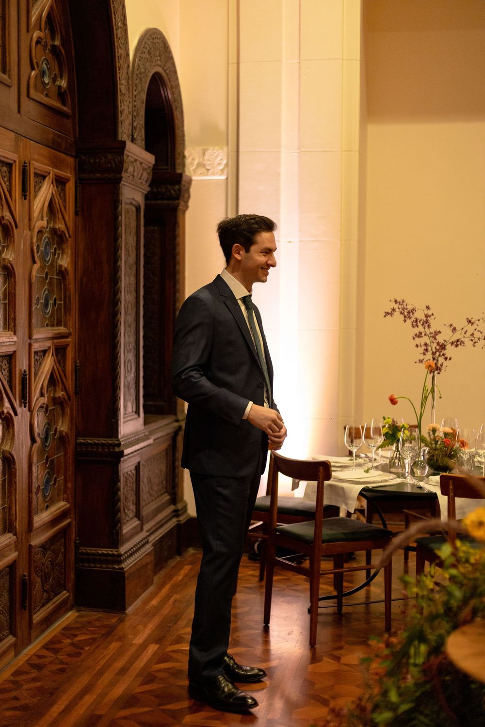 A well-dressed man in a dark suit stands in an elegant room with ornate wooden archways and a dining table setup.