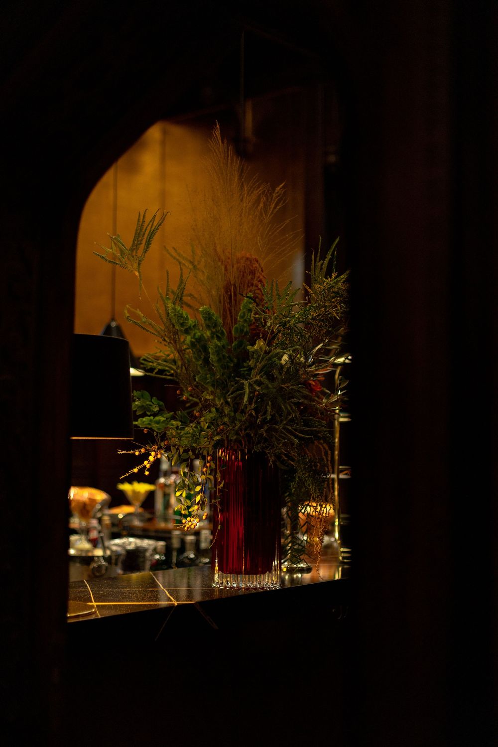 A floral arrangement in a red vase viewed through a dark window frame with warm lighting in the background.