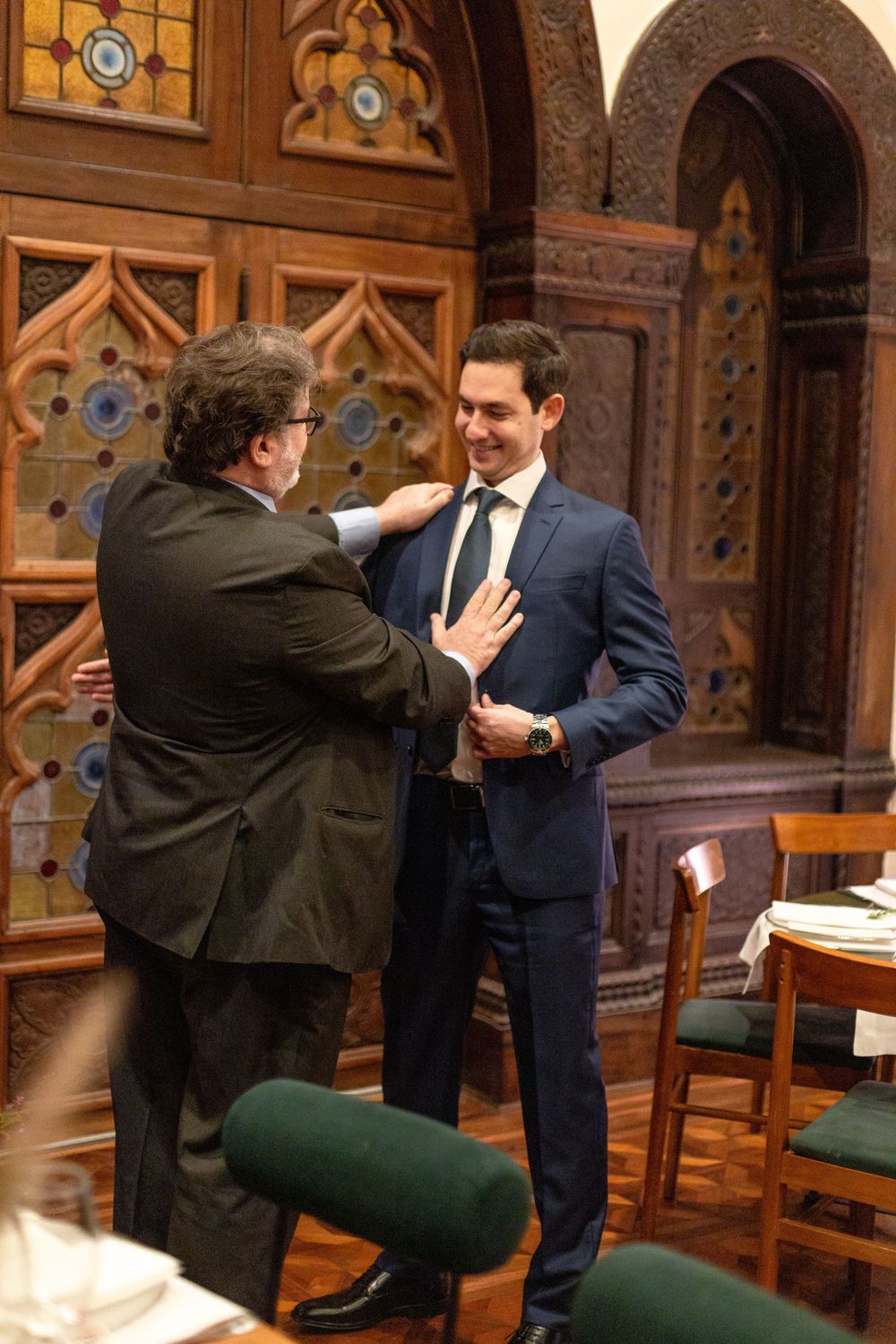 Two men in suits engage in an animated conversation inside an ornate wood-paneled room with stained glass windows.