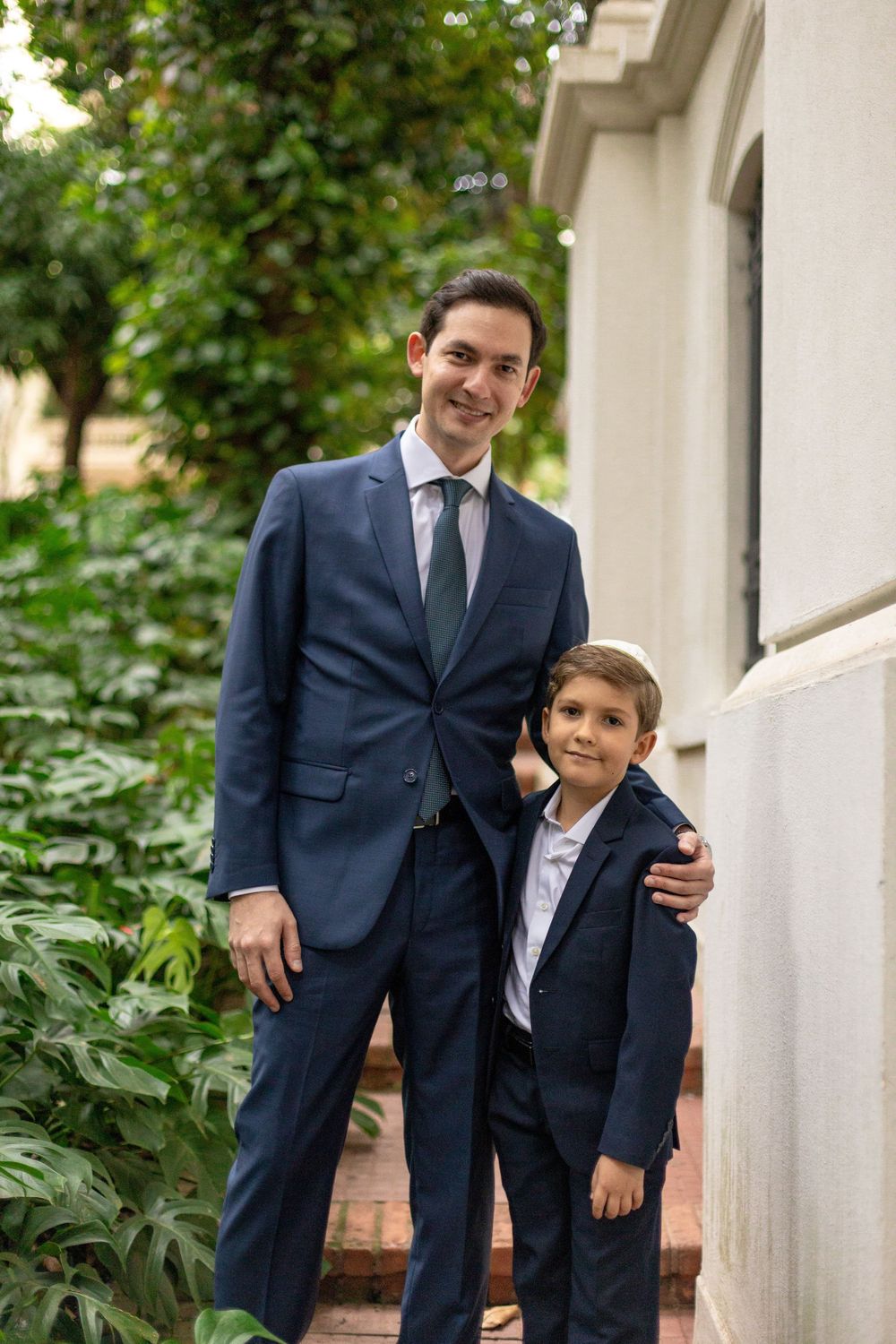 Two people in matching navy suits stand together outdoors by a white building with greenery in the background.
