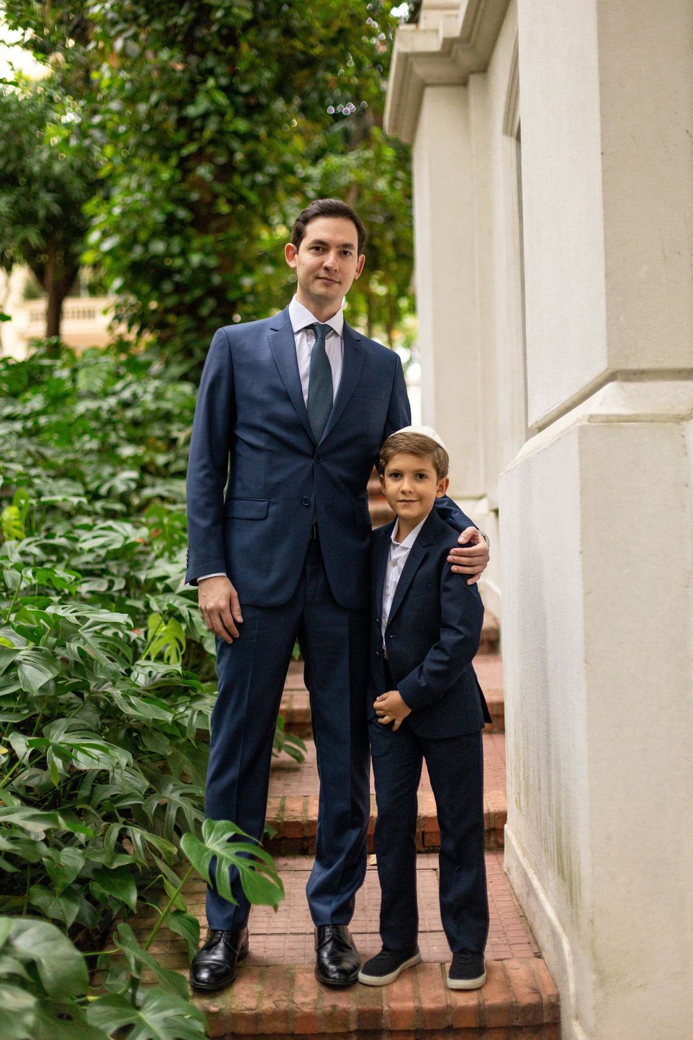 Two people in matching navy suits stand together between white columns in an outdoor garden setting.