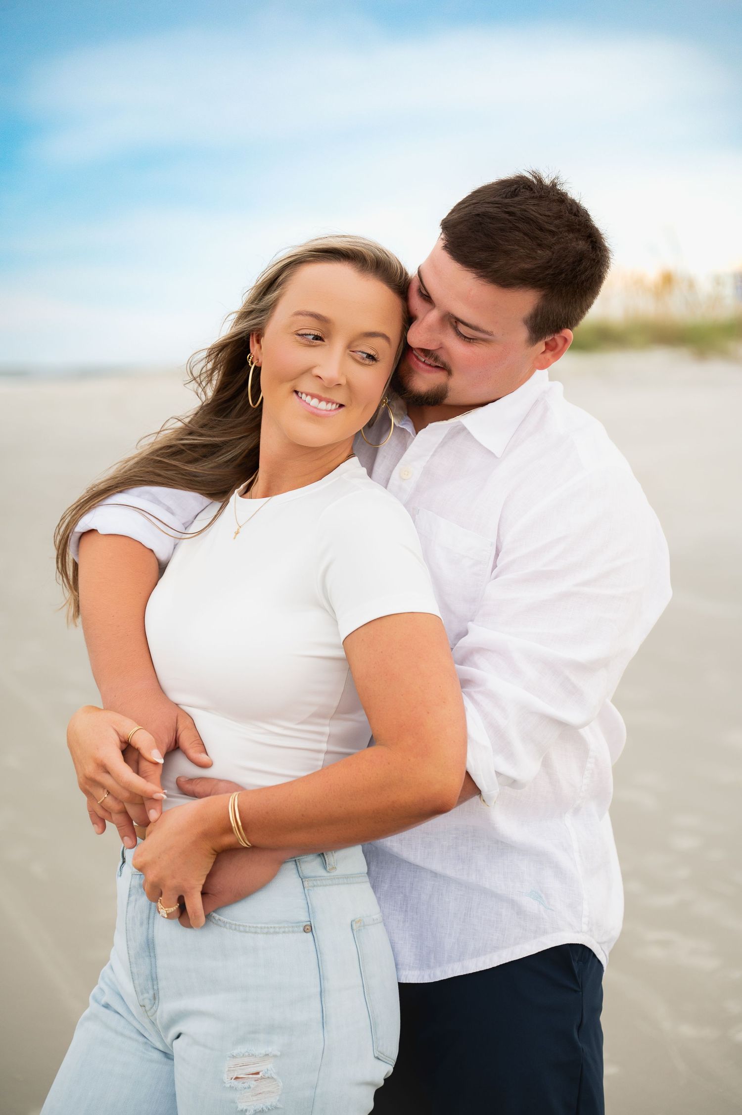 Couple embracing on beach with ocean waves and blue sky in background.