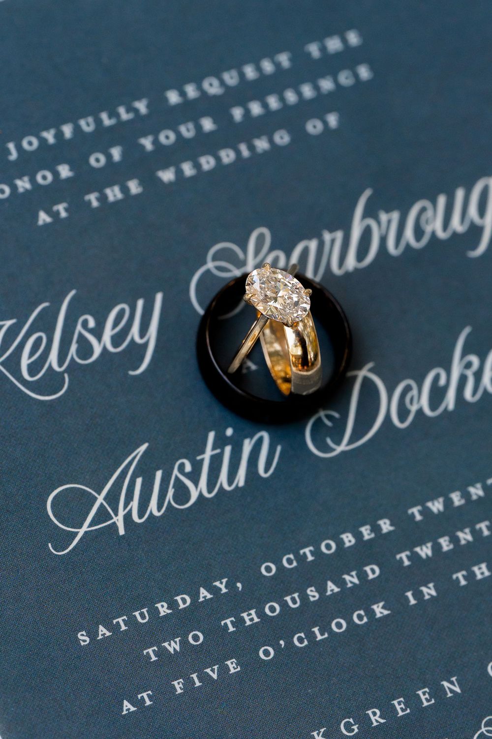 Diamond engagement ring and wedding band displayed on navy blue invitation with calligraphy.