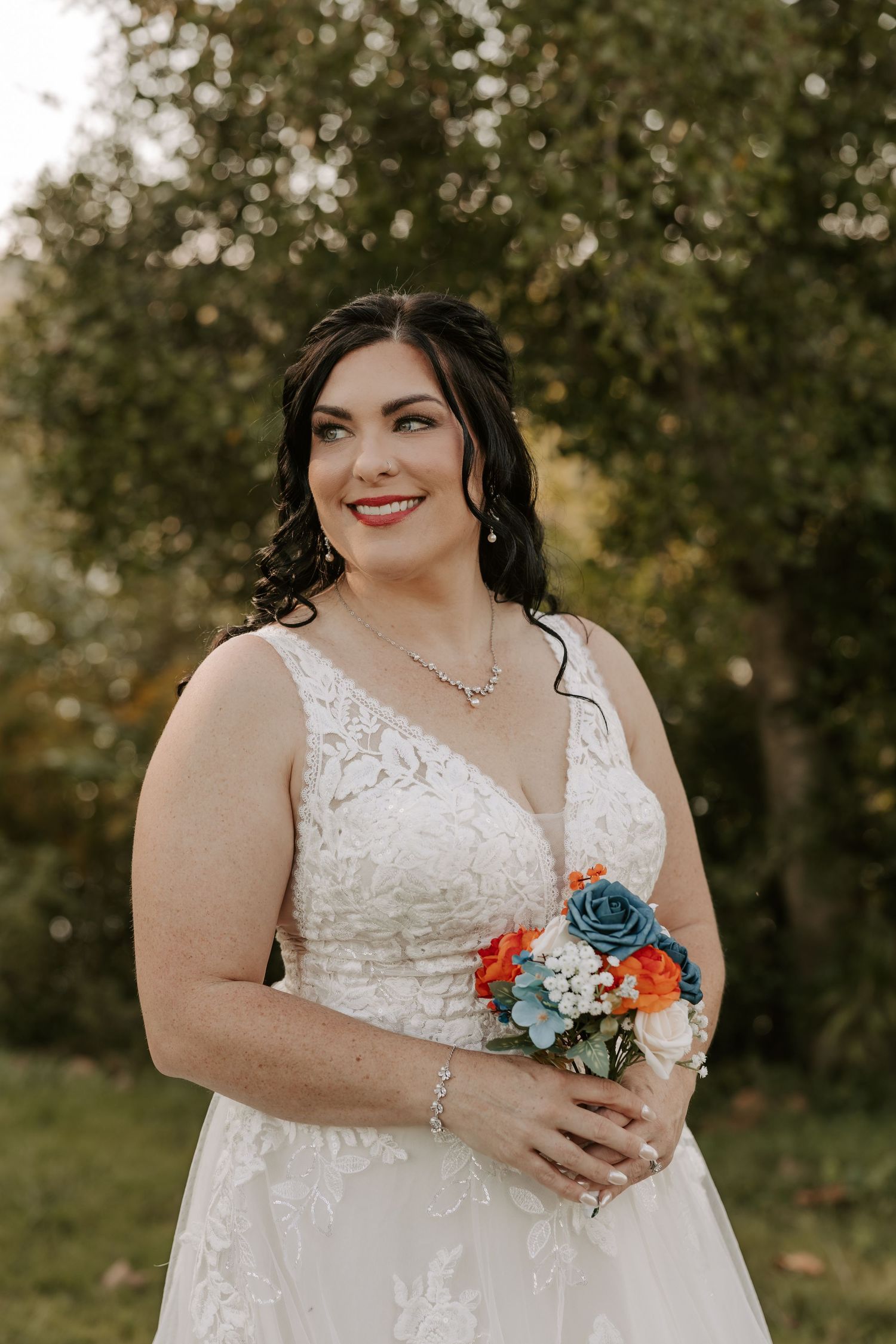 A bride in a white lace wedding dress holds a vibrant coral and blue floral bouquet in an outdoor garden setting.