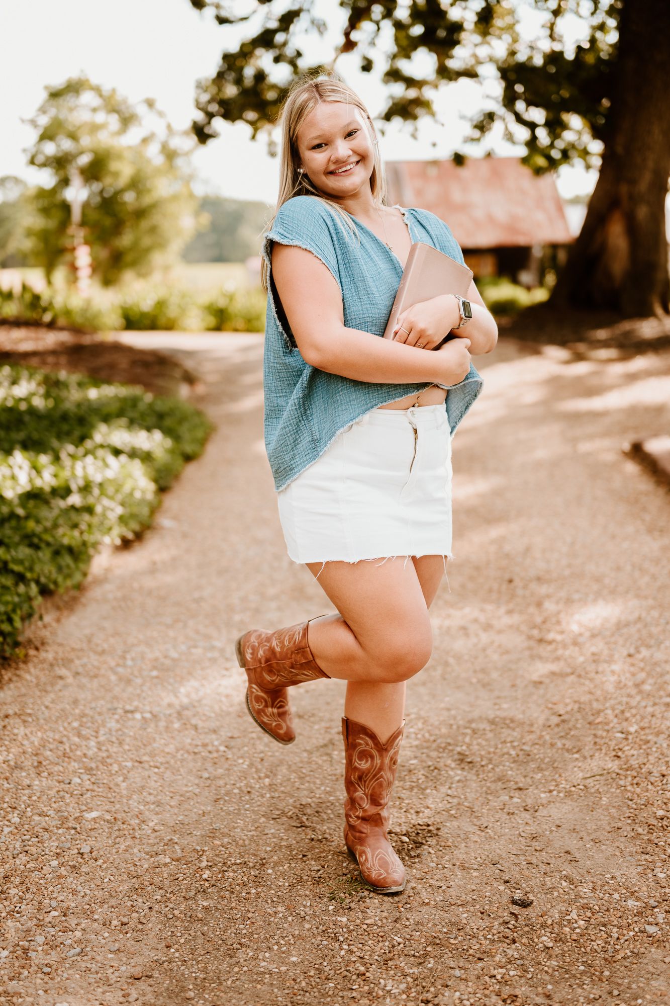 A person in a blue top and brown cowboy boots dances playfully on a dirt path in a sunny outdoor setting.
