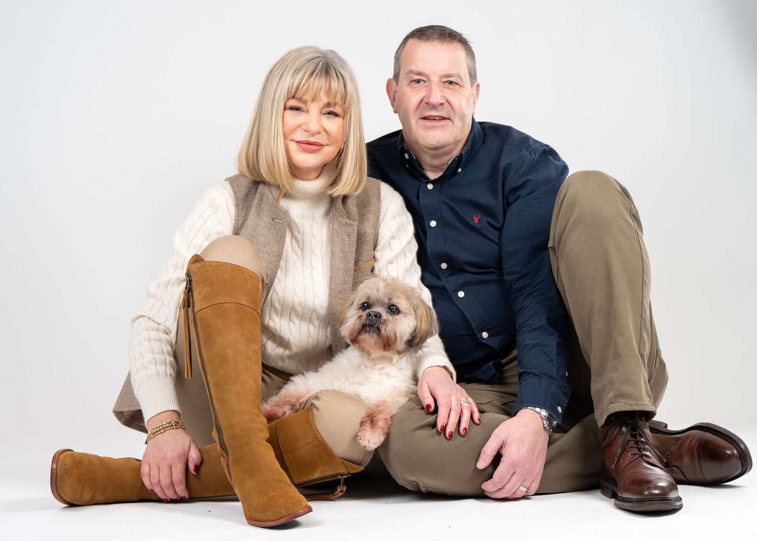 A couple in casual attire sits on a white background with their small fluffy dog in a series of relaxed poses.