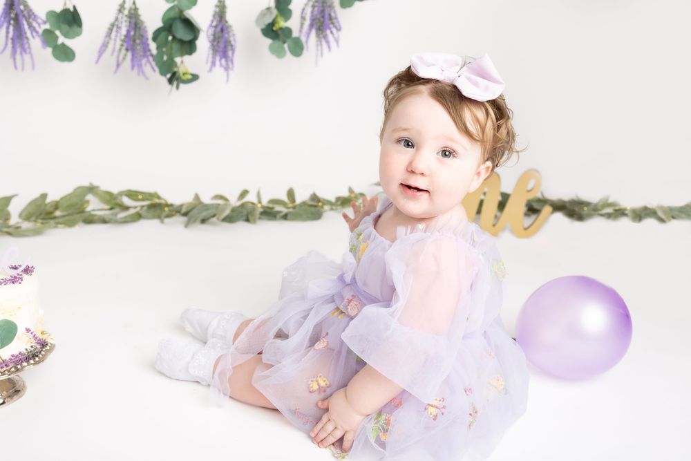 A baby in a purple tutu dress sits among purple balloons and floral decorations during a photo session.