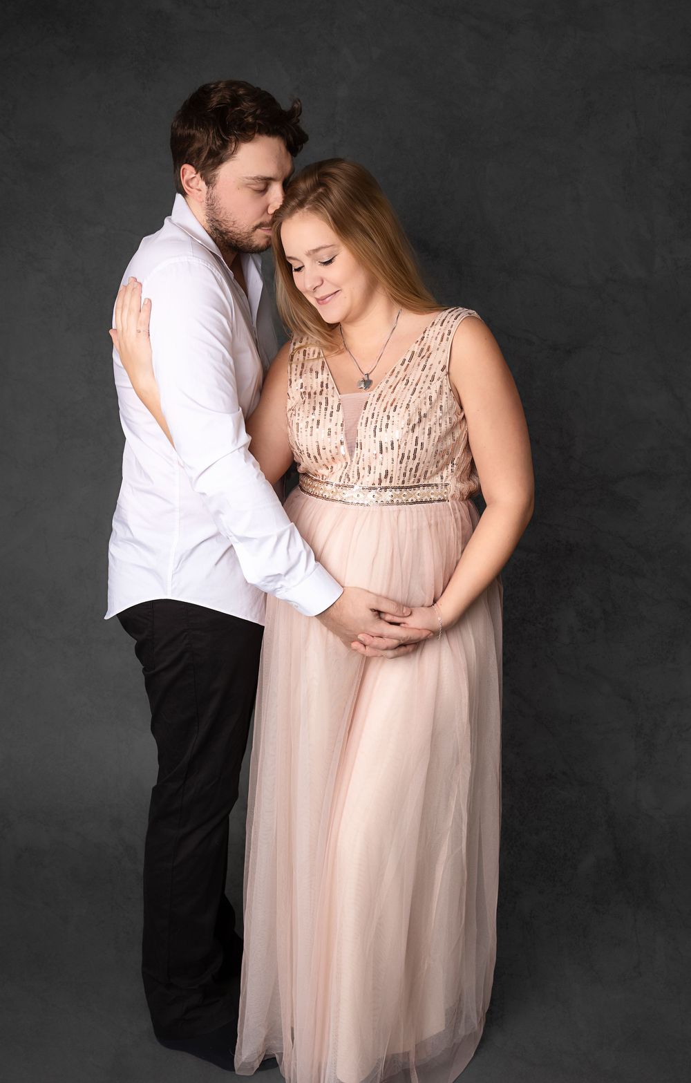 A couple shares an intimate maternity photo moment against a dark backdrop in a flowing blush gown and formal attire.