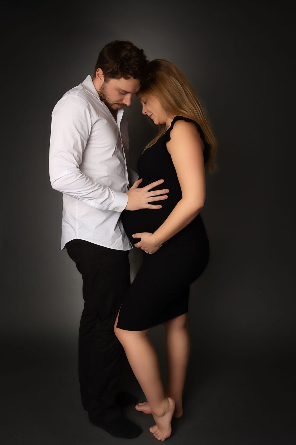 A couple shares an intimate moment during a maternity photoshoot against a dark backdrop.