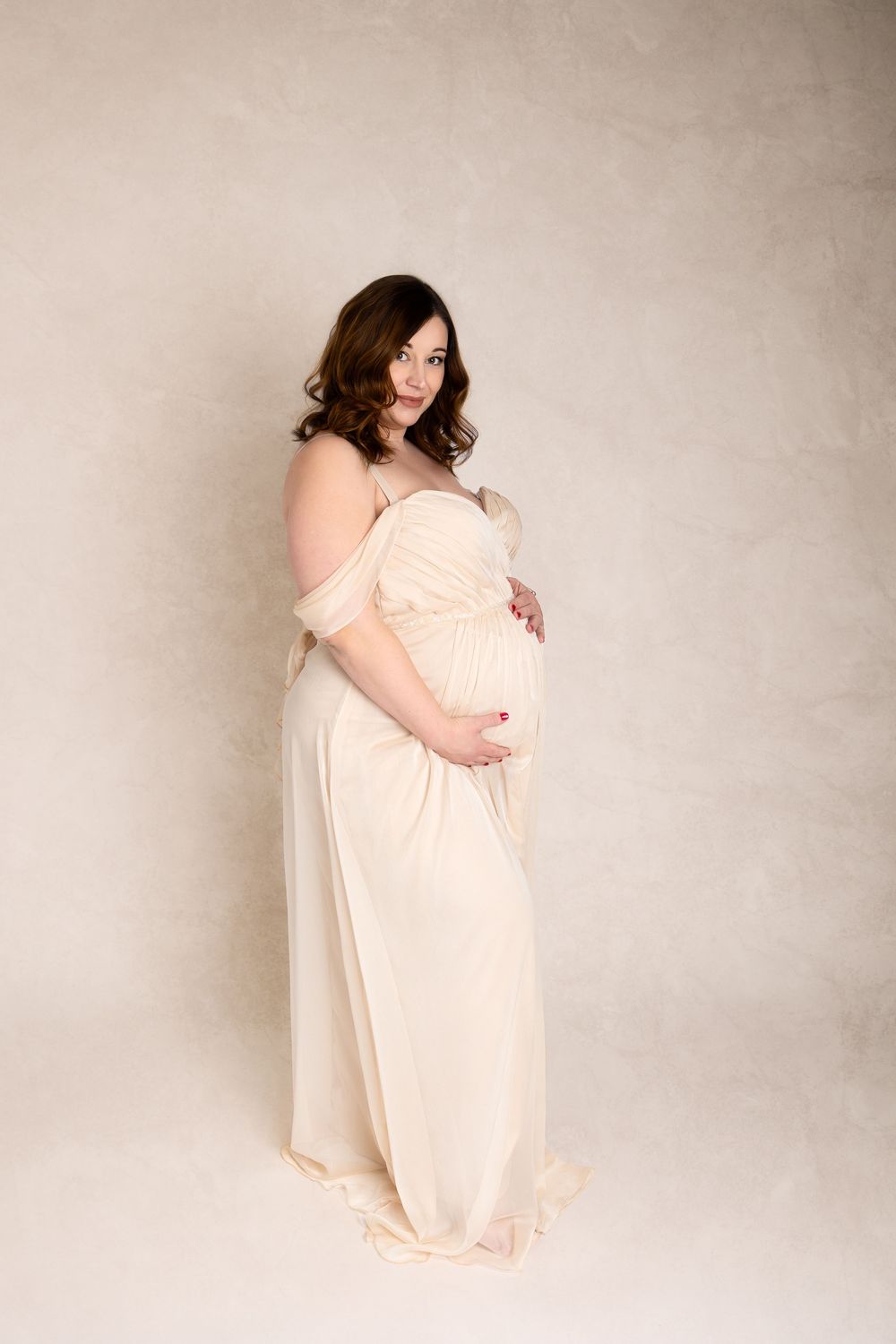 A pregnant woman in a flowing off-shoulder peach gown poses against a neutral backdrop during a maternity photoshoot.