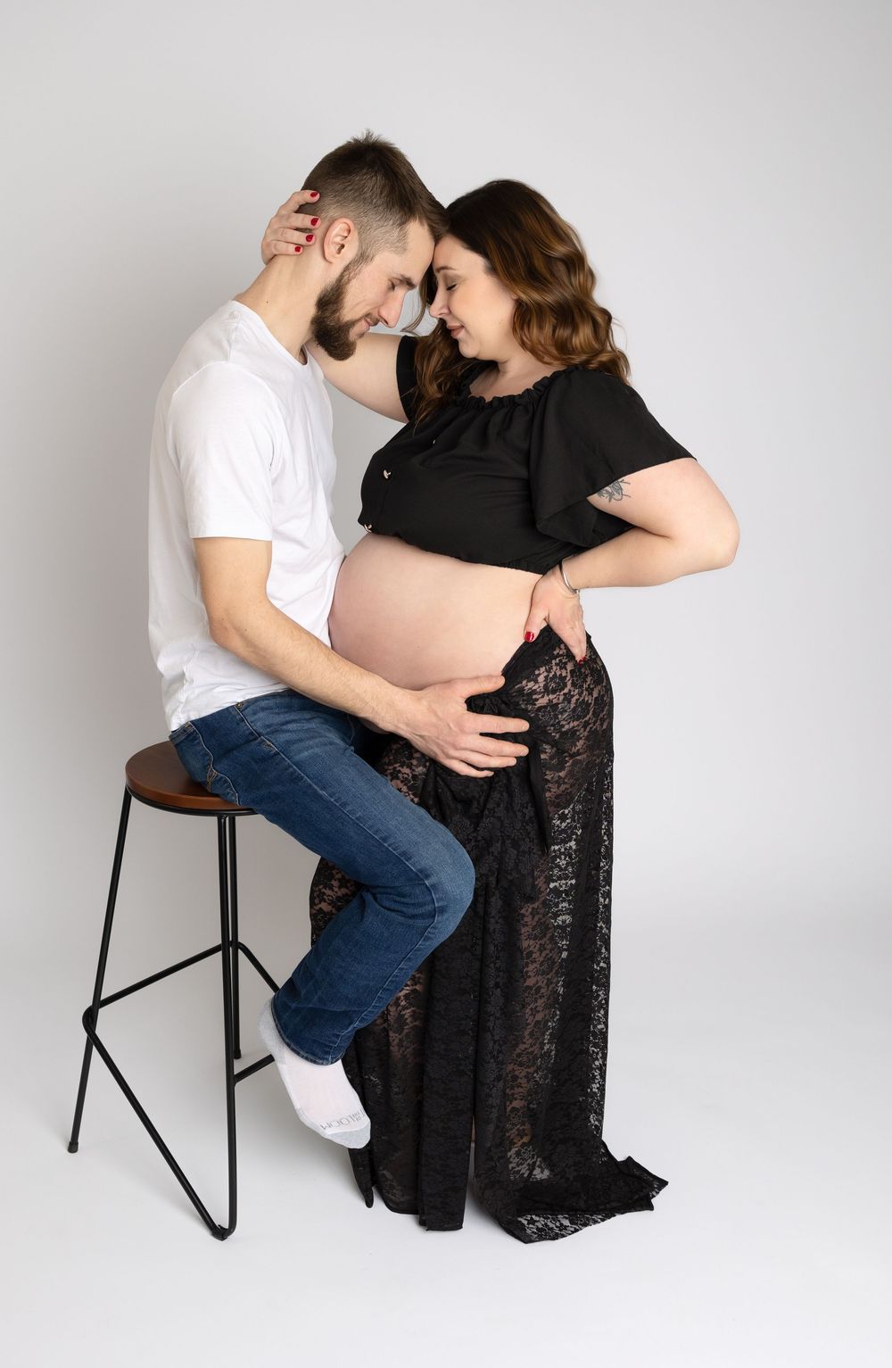 A couple in white and black outfits share an intimate moment while sitting on a stool against a light gray backdrop.