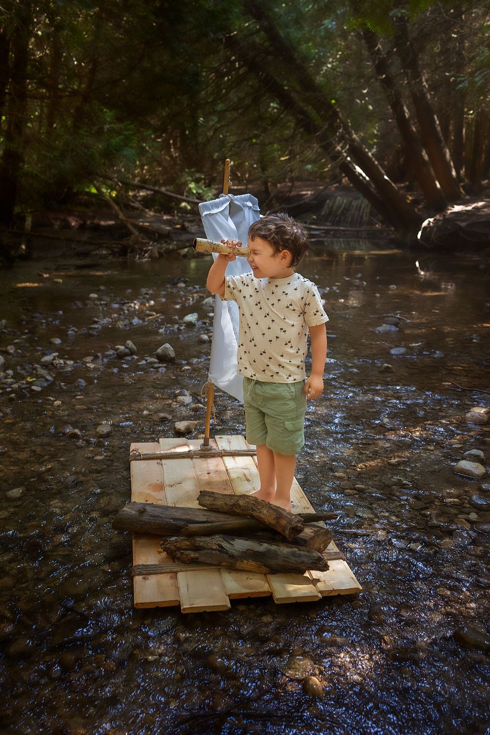 Child in light shirt and green shorts stands on a wooden raft in a shallow creek while drinking water.