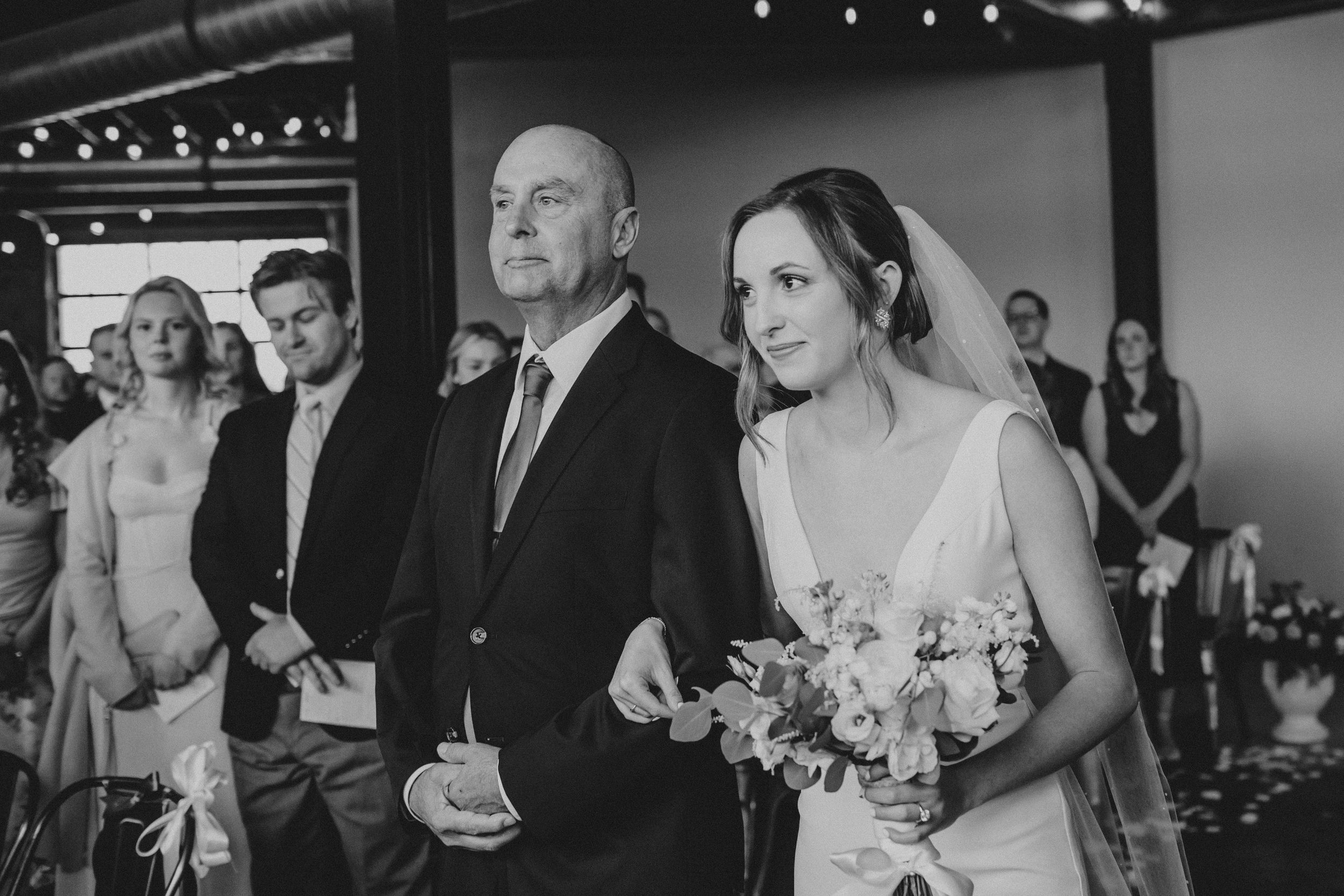 Black and white photo of a bride walking down the aisle with her escort at an indoor wedding ceremony.