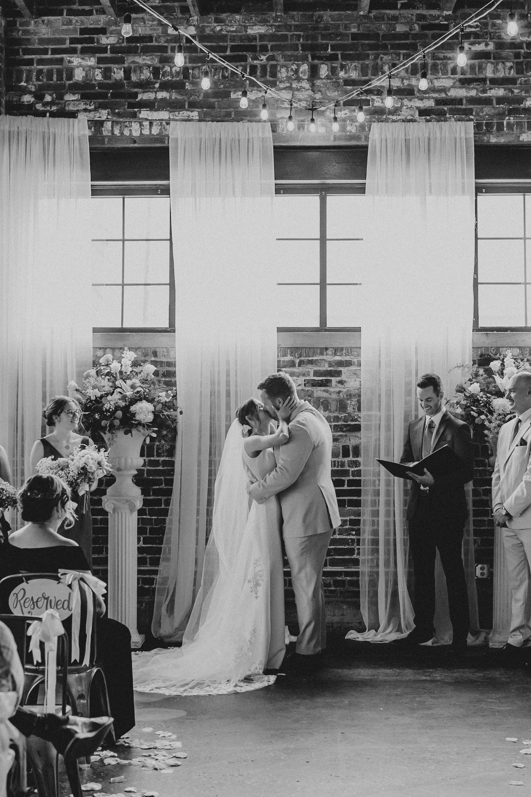 A black and white wedding ceremony photo showing a couple embracing at the altar with white curtains and brick walls.