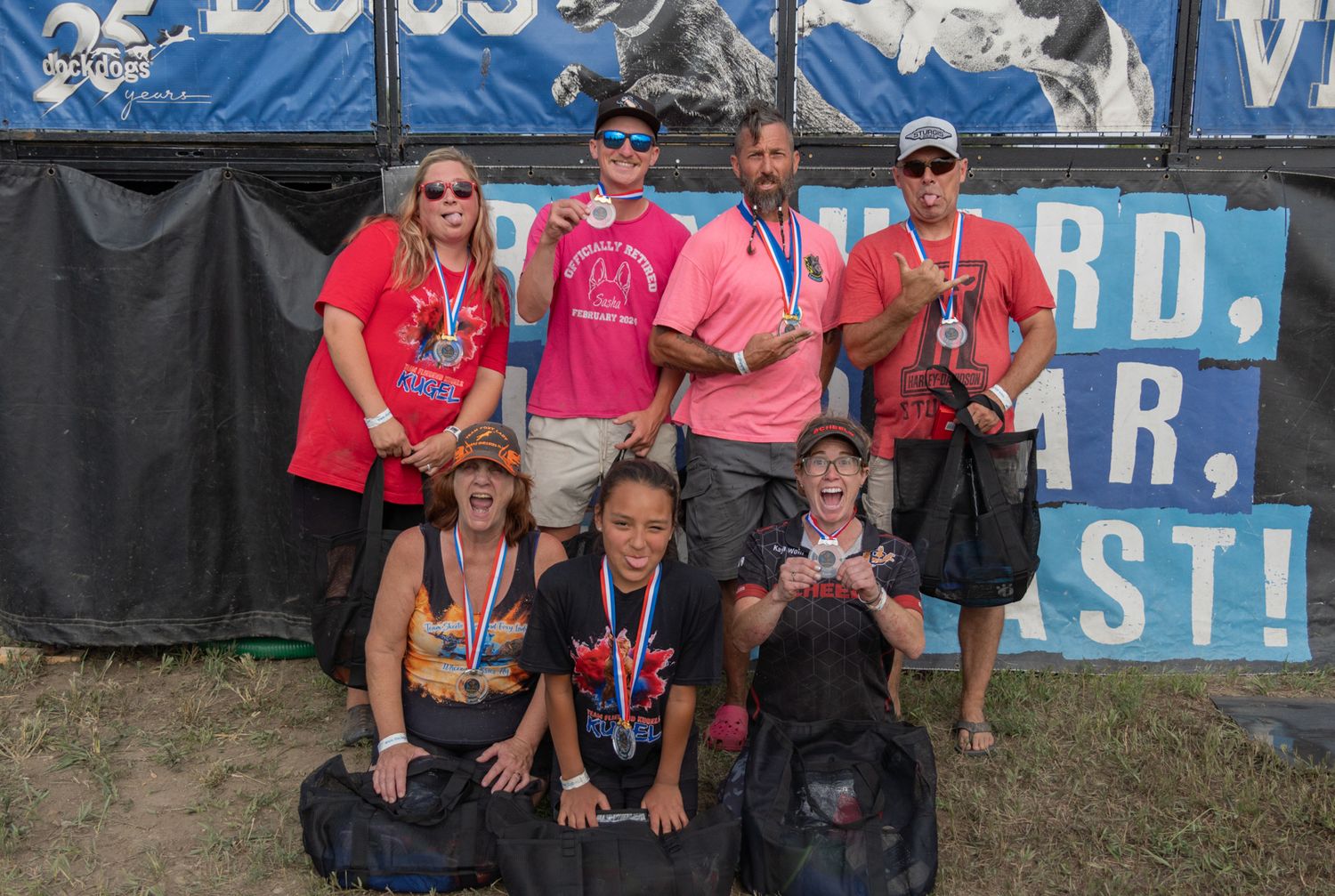 Group of adults and children posing with medals at an outdoor event in front of a motivational banner.