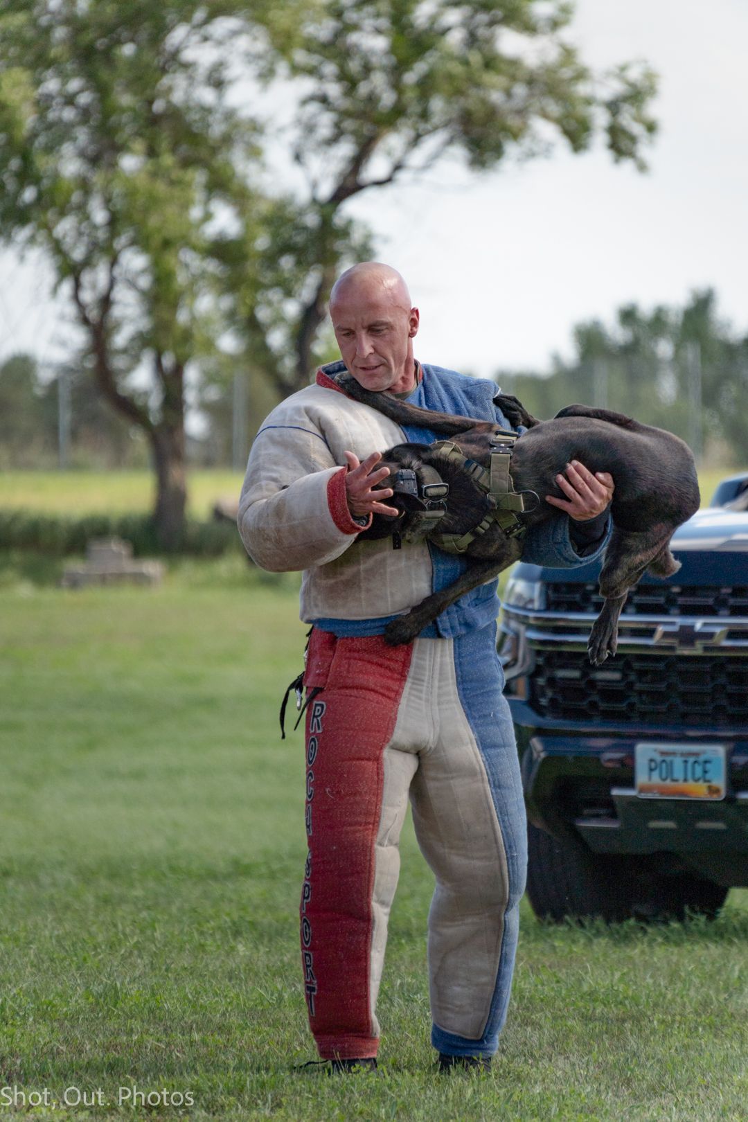 Person in protective training gear holding and carrying a large dog during an outdoor training exercise.