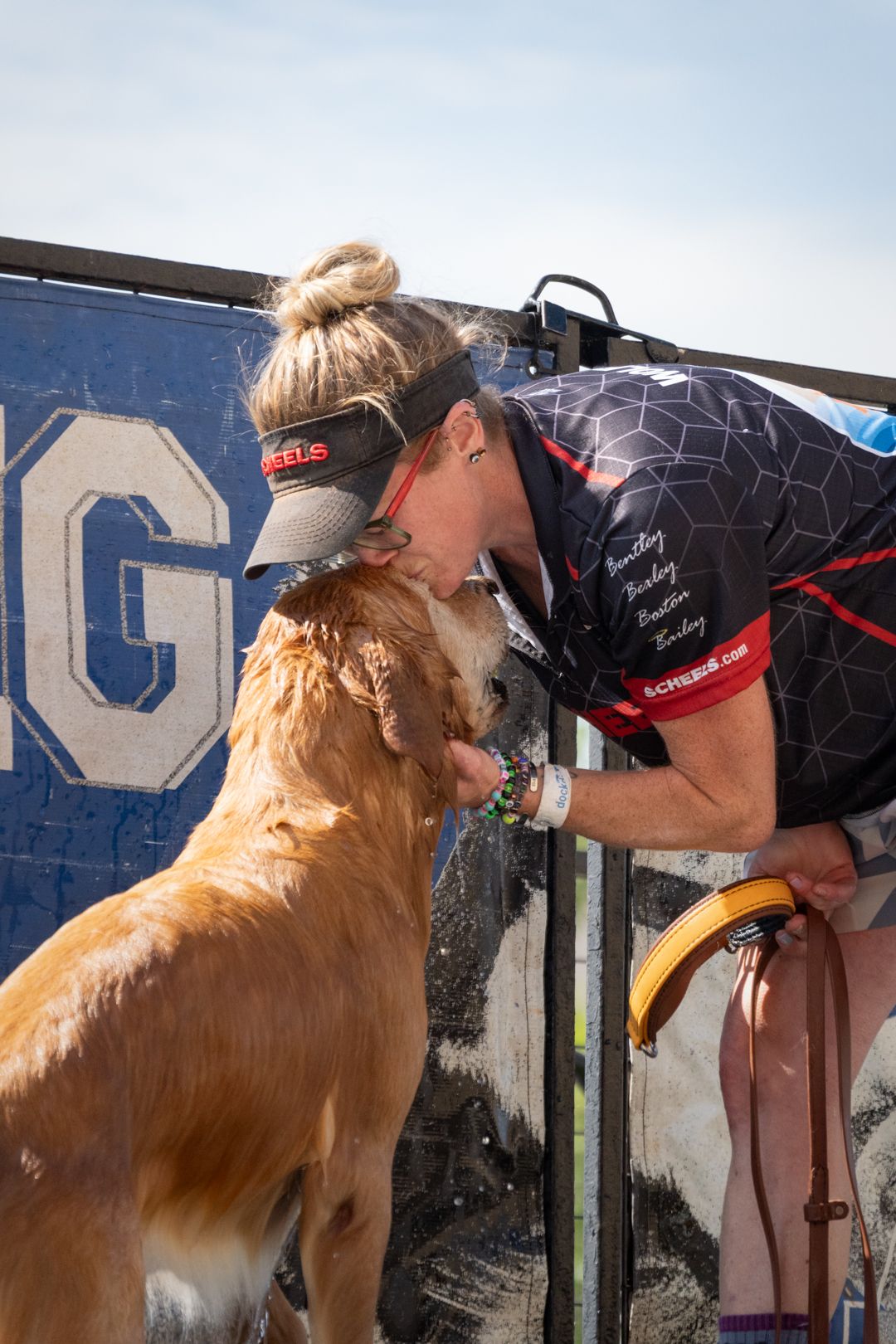 Woman in black jersey and cap affectionately nuzzling a golden retriever dog outdoors at an event.