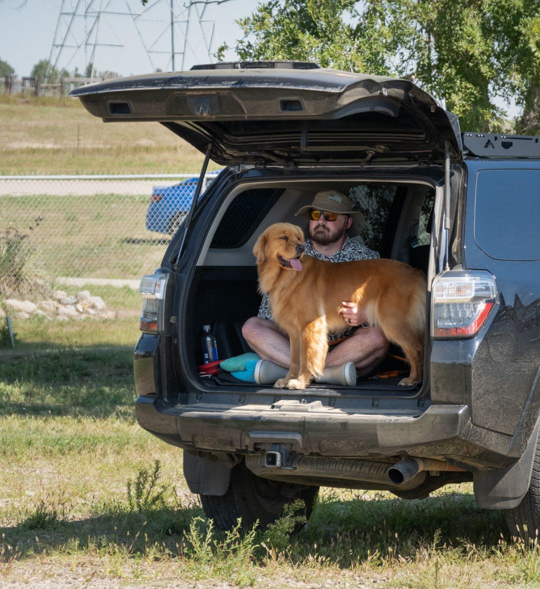 Man in hat sitting in open SUV trunk with golden retriever dog parked on grassy field near trees.