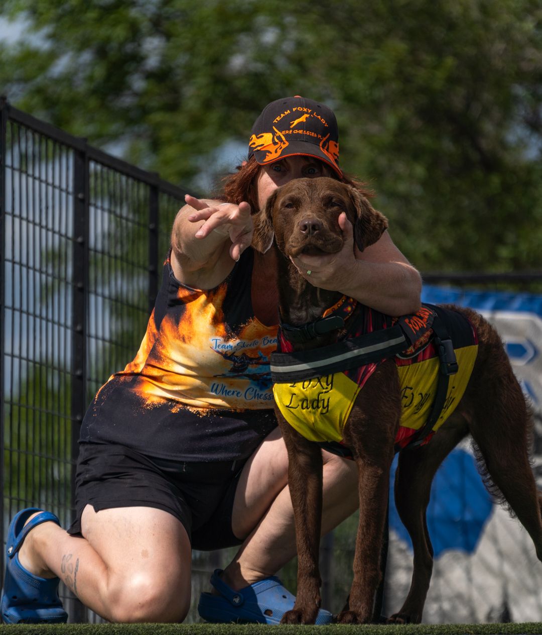 A chocolate Labrador puppy wearing a colorful service vest sits with someone outdoors near a fence.