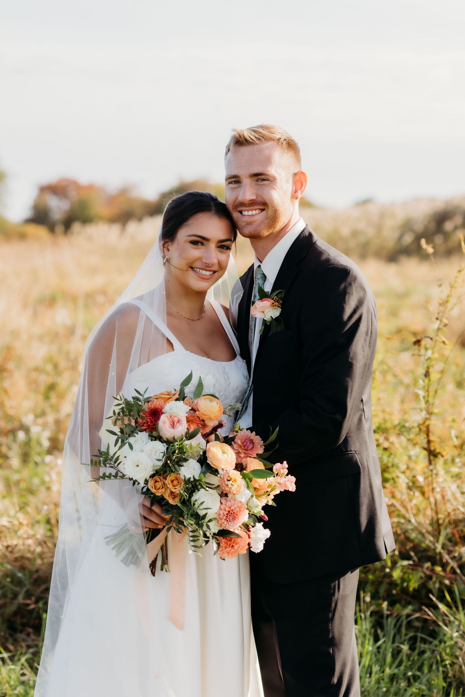 A couple poses together in a golden field at sunset with the bride holding a peach and coral floral bouquet.