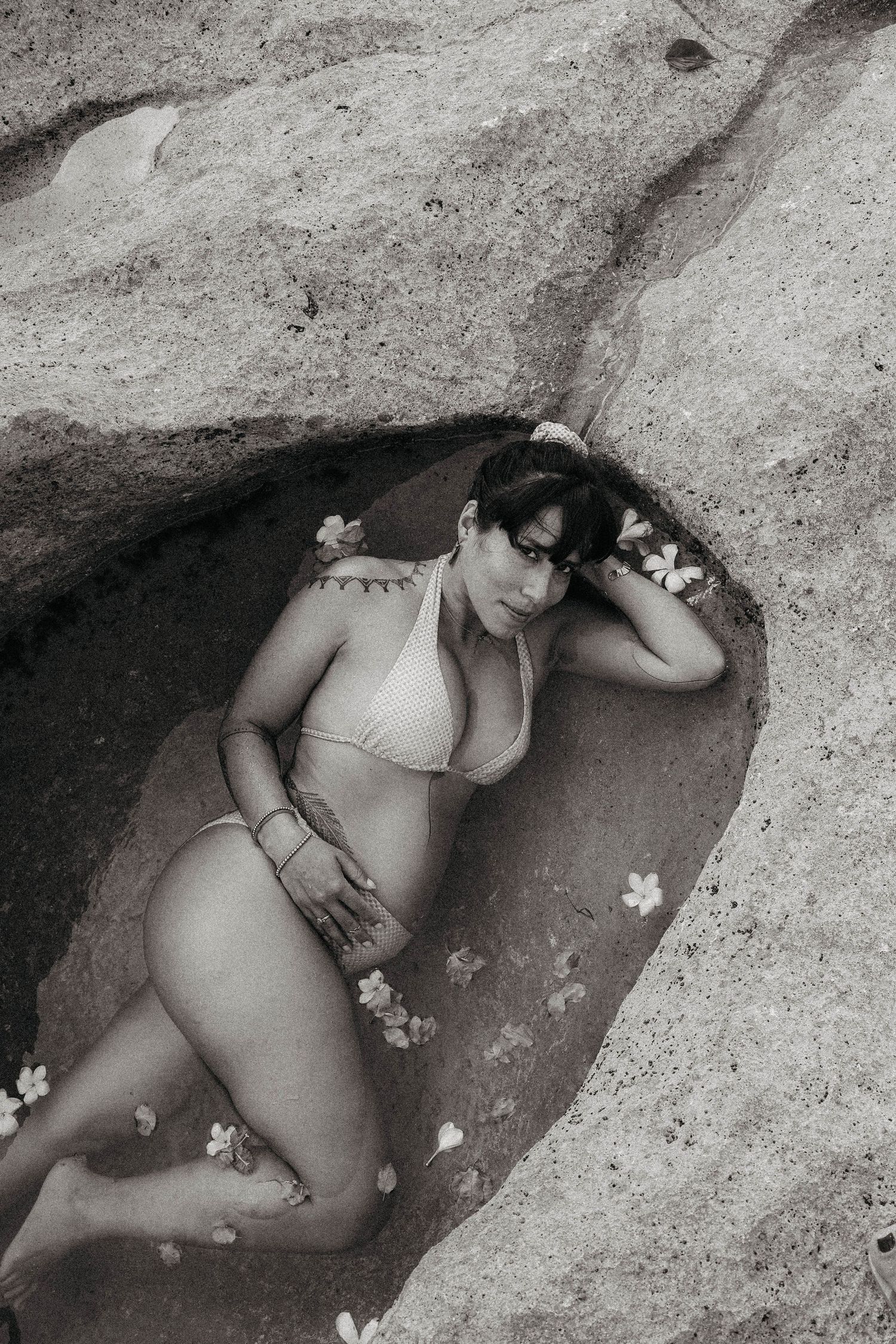 Black and white photo of a woman in white bikini posing in a rocky coastal cave with flowers.