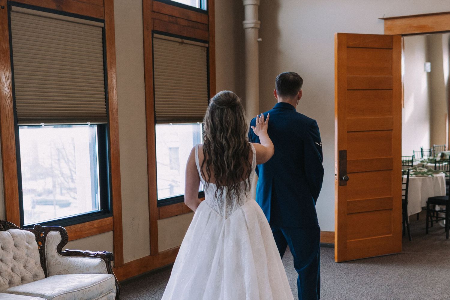 A couple in wedding attire stands by large windows in a rustic room.