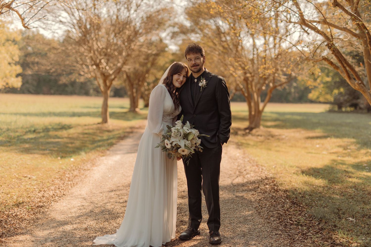 Couple stands peacefully on a sunlit dirt path surrounded by golden autumn trees, bride holding a white bouquet.