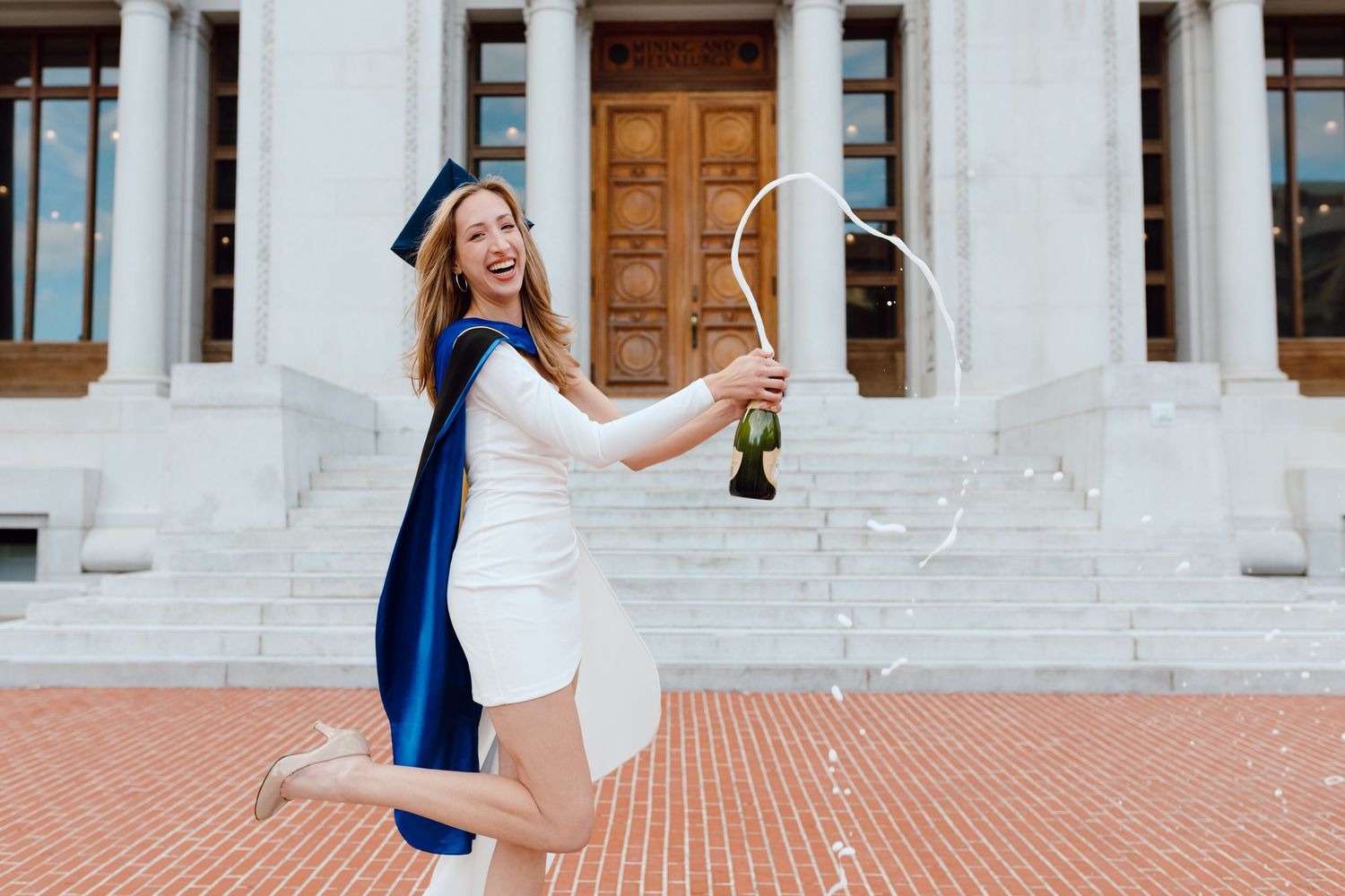 UC Berkeley graduate smiles and laughs as she pops and sprays champagne outside of Hearst Mining Building on campus for her grad photoshoot.