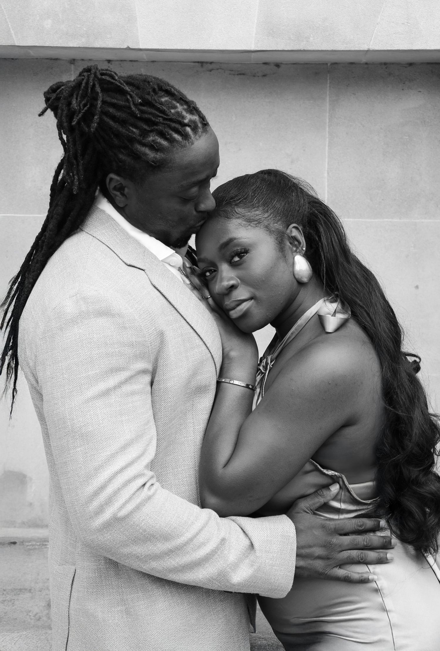 A couple in elegant white wedding attire poses together in front of a lush green hedge wall.