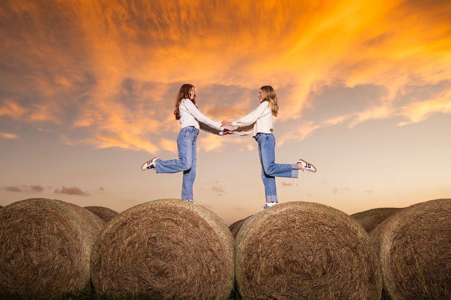 Silhouettes posing on hay bales against dramatic orange sunset sky in rural field.