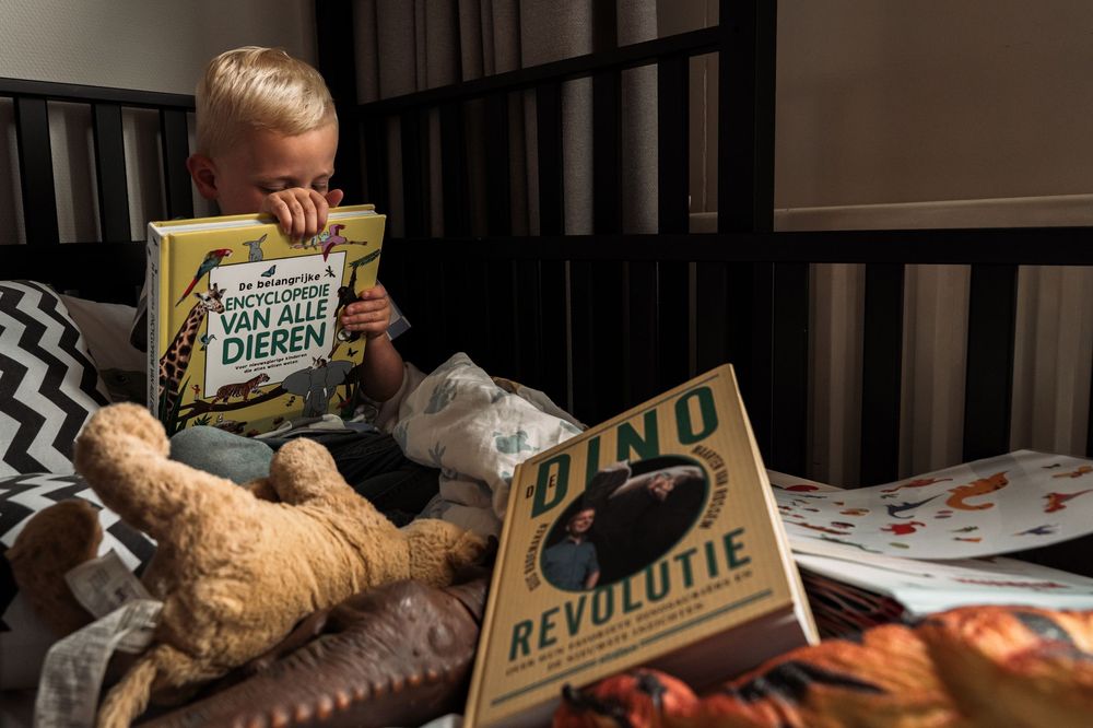 A child reading books in a crib with a stuffed animal companion.