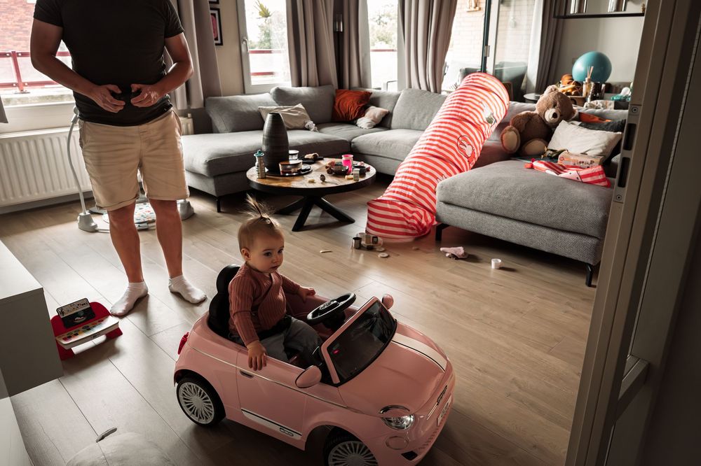 A child rides in a pink toy car while adults relax on a gray couch in the background.
