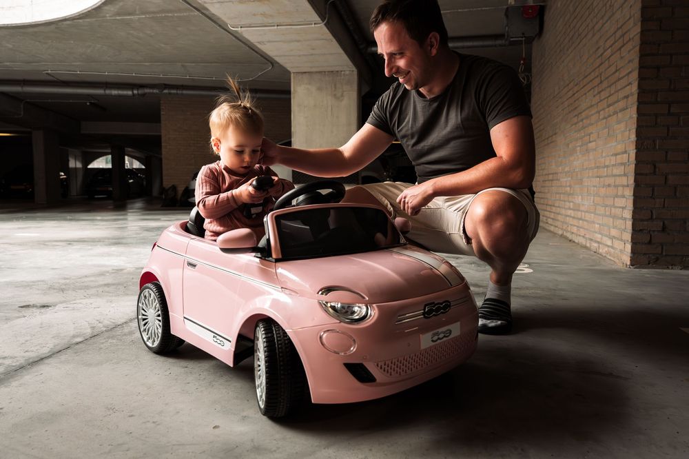 A parent helps guide a small child driving a pink toy car in a parking garage.