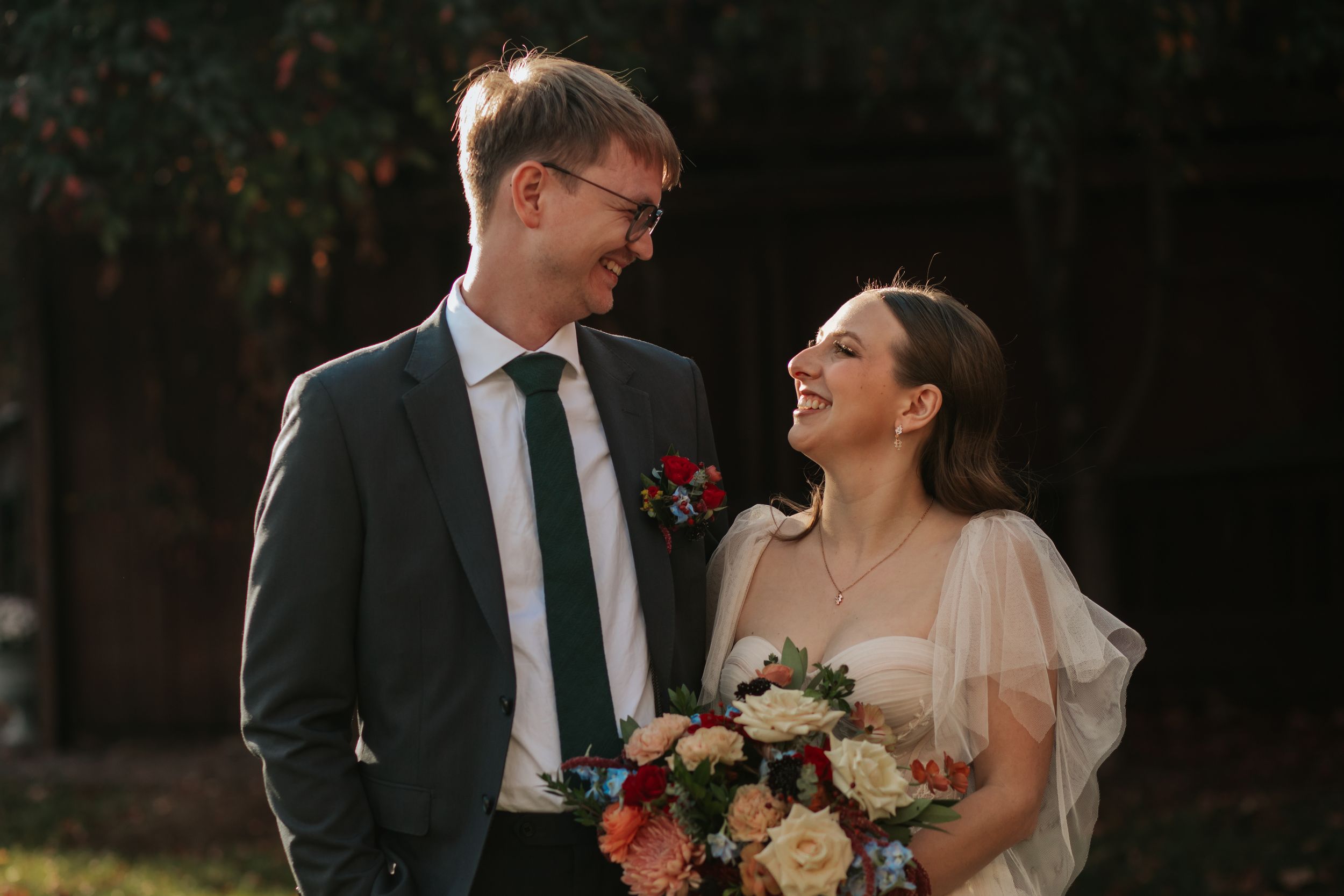 A couple in wedding attire laughs together in a romantic outdoor setting with soft lighting and natural greenery.