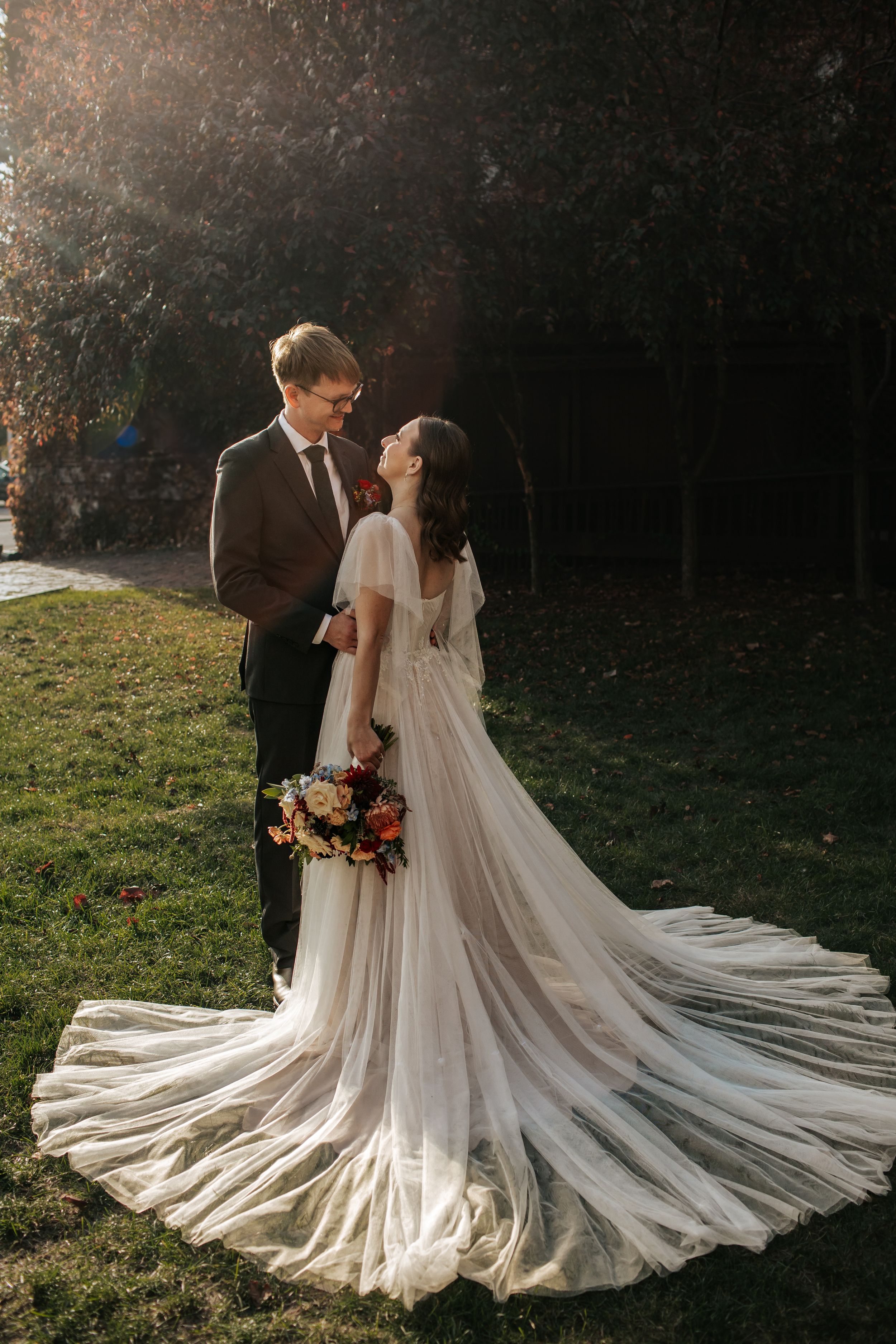 Elegant wedding couple poses in nature with bride's flowing cathedral veil creating dramatic circular shape at sunset.