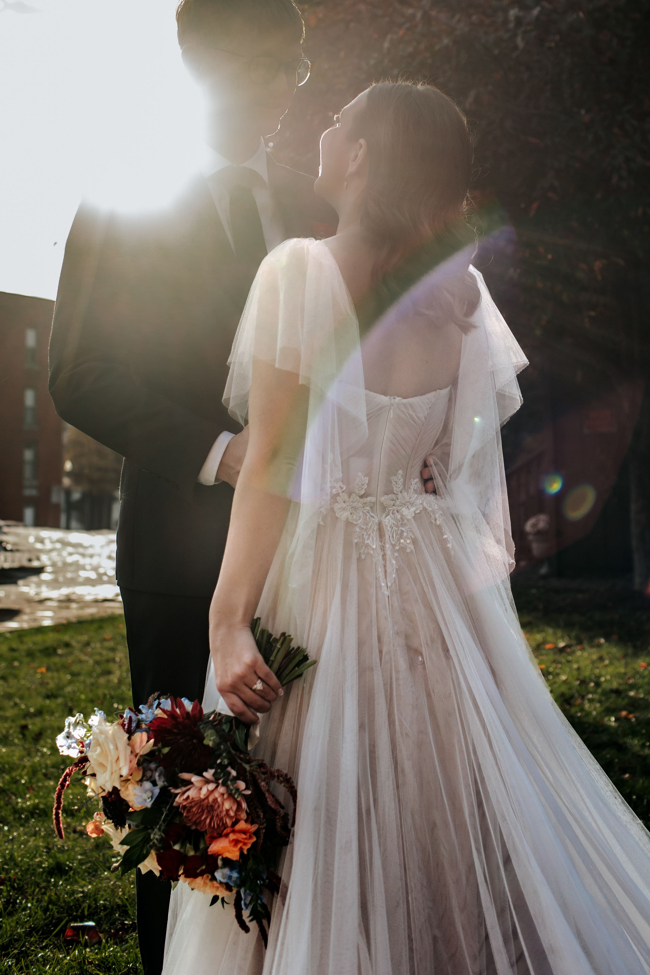 Romantic backlit wedding photo of couple in formal attire with flowing white dress and floral bouquet at sunset.
