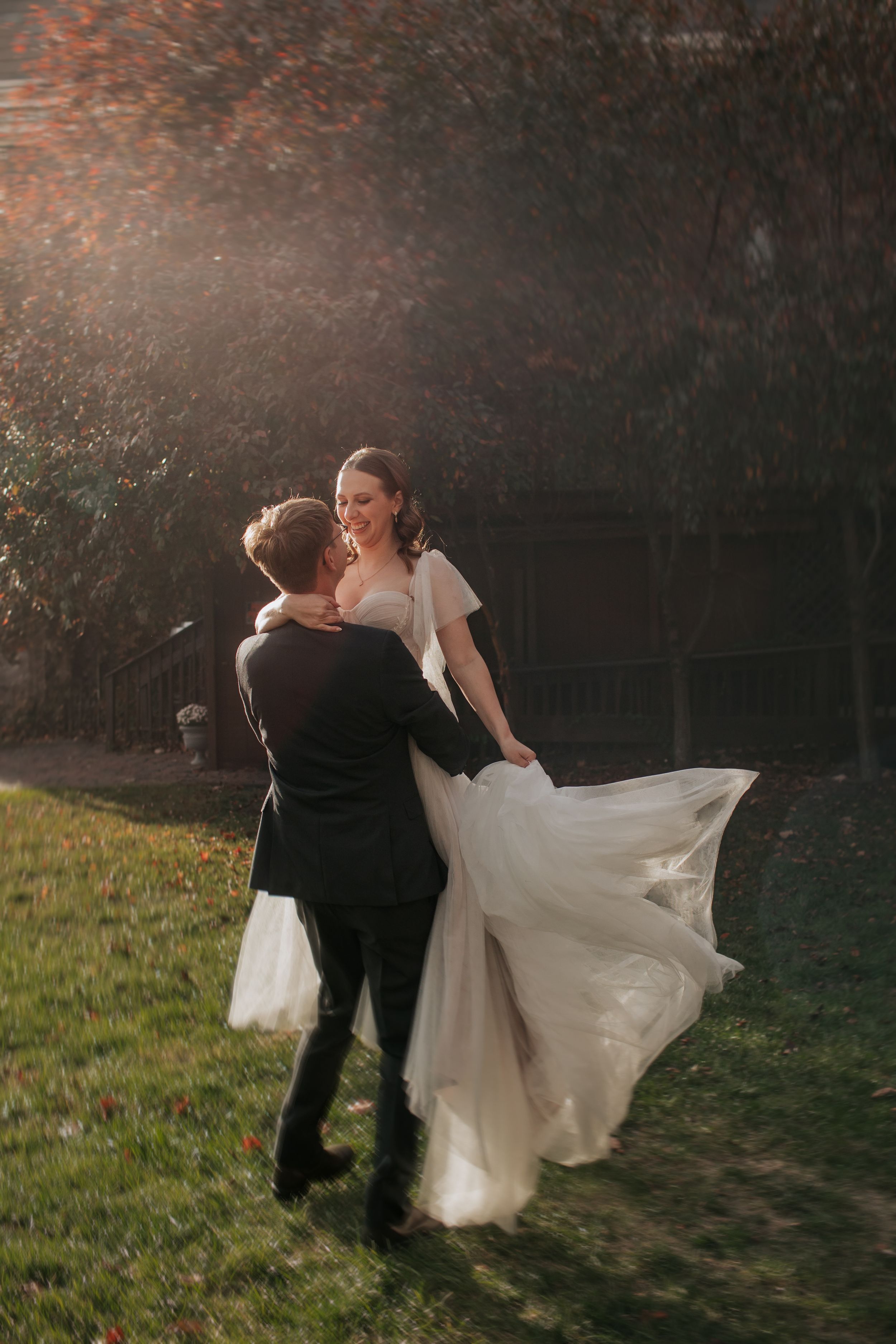 A bride and groom dance together in a romantic outdoor setting with golden sunlight filtering through autumn trees.