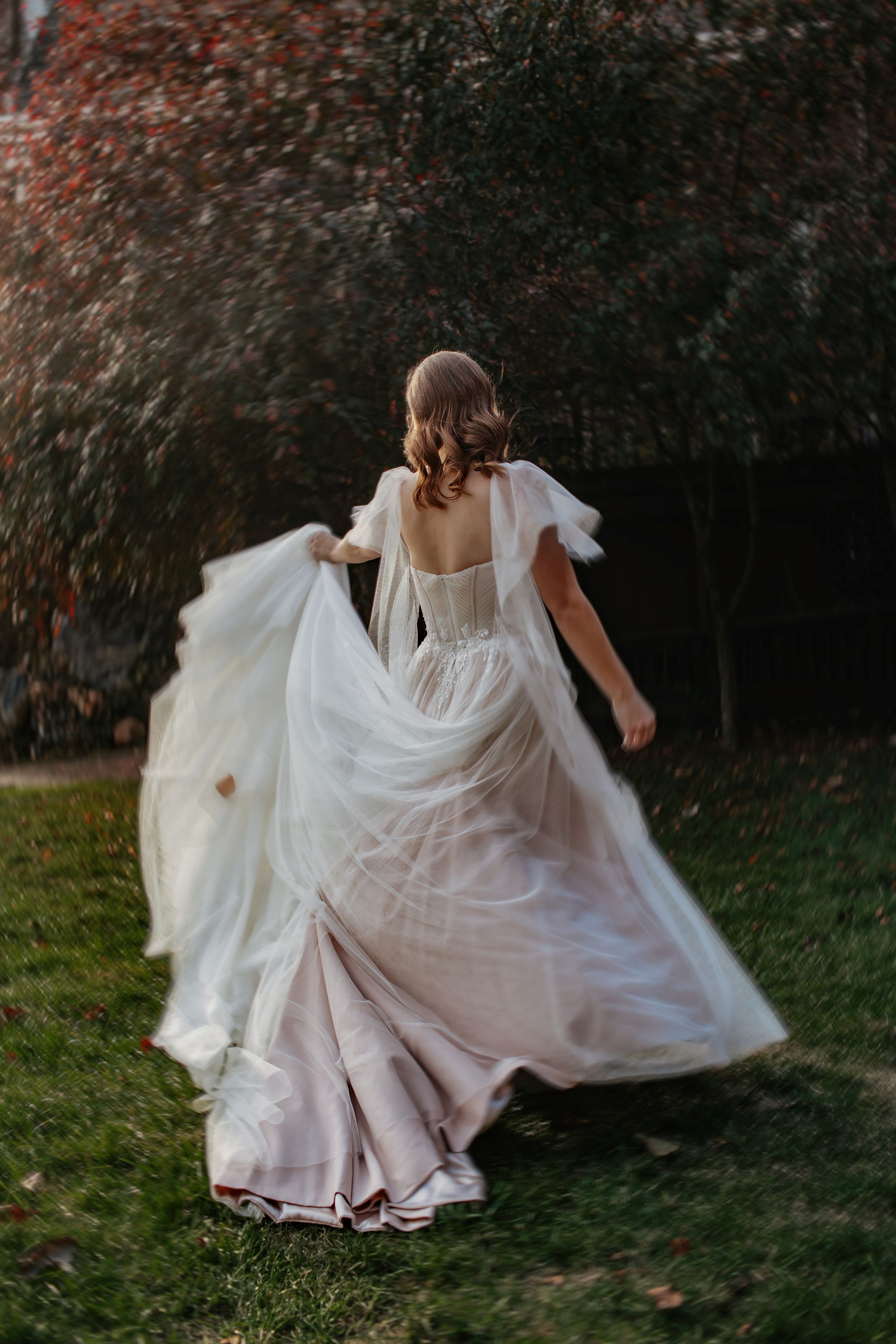 A person in a flowing white dress twirls gracefully in a garden setting with autumn foliage in the background.