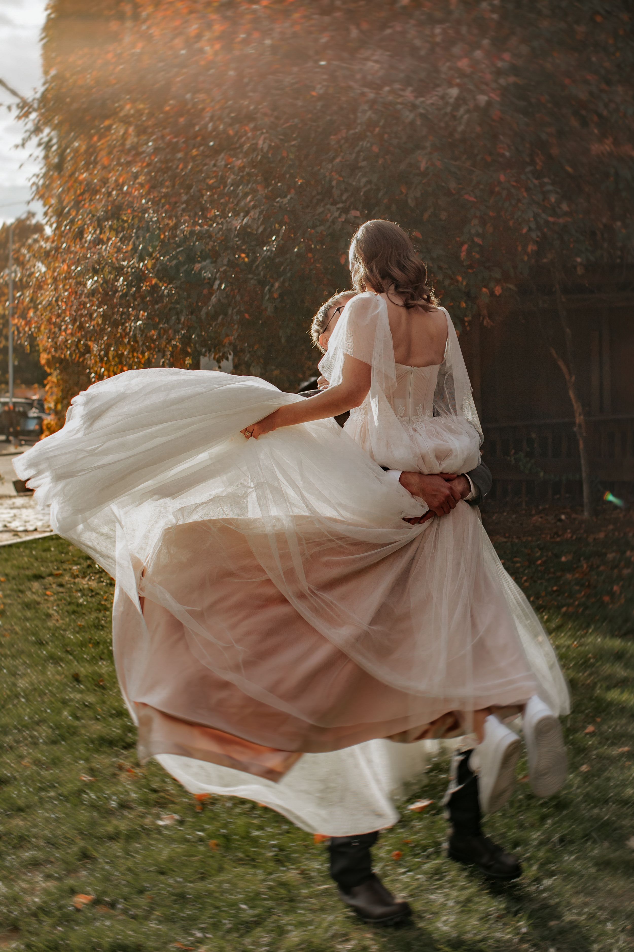 A couple in wedding attire dances and twirls in a romantic autumn setting as the bride's white dress flows in the breeze.
