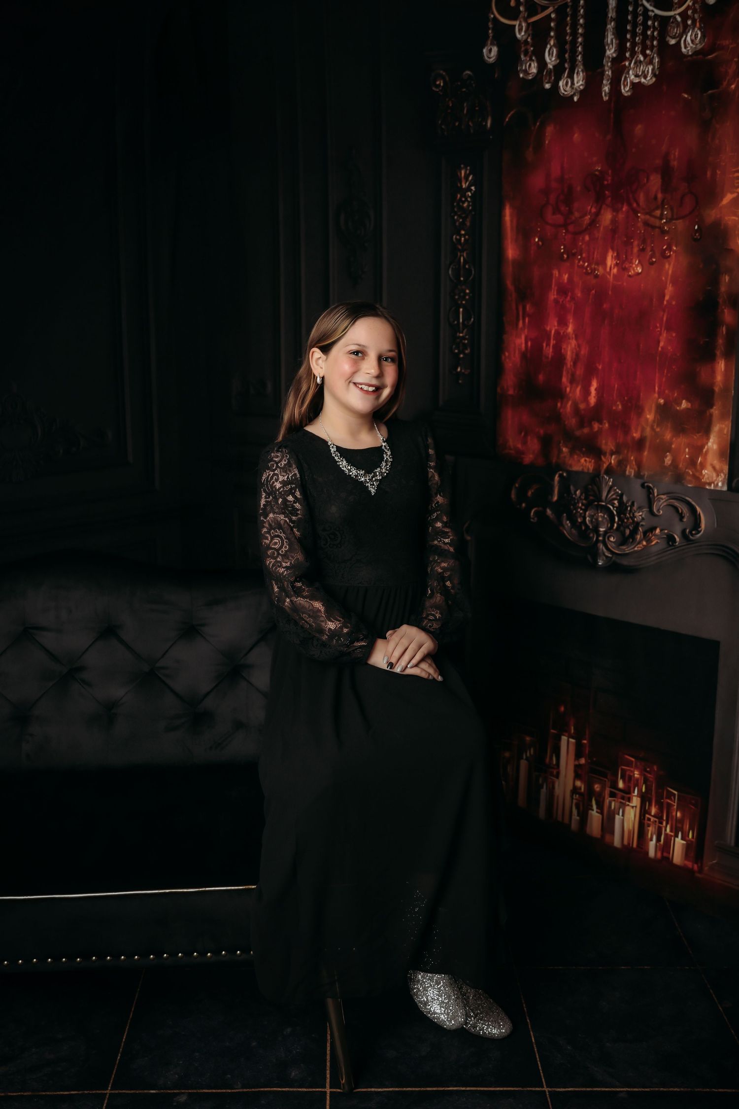 A person wearing a black lace dress poses on a dark velvet couch against a dramatic red and black backdrop.
