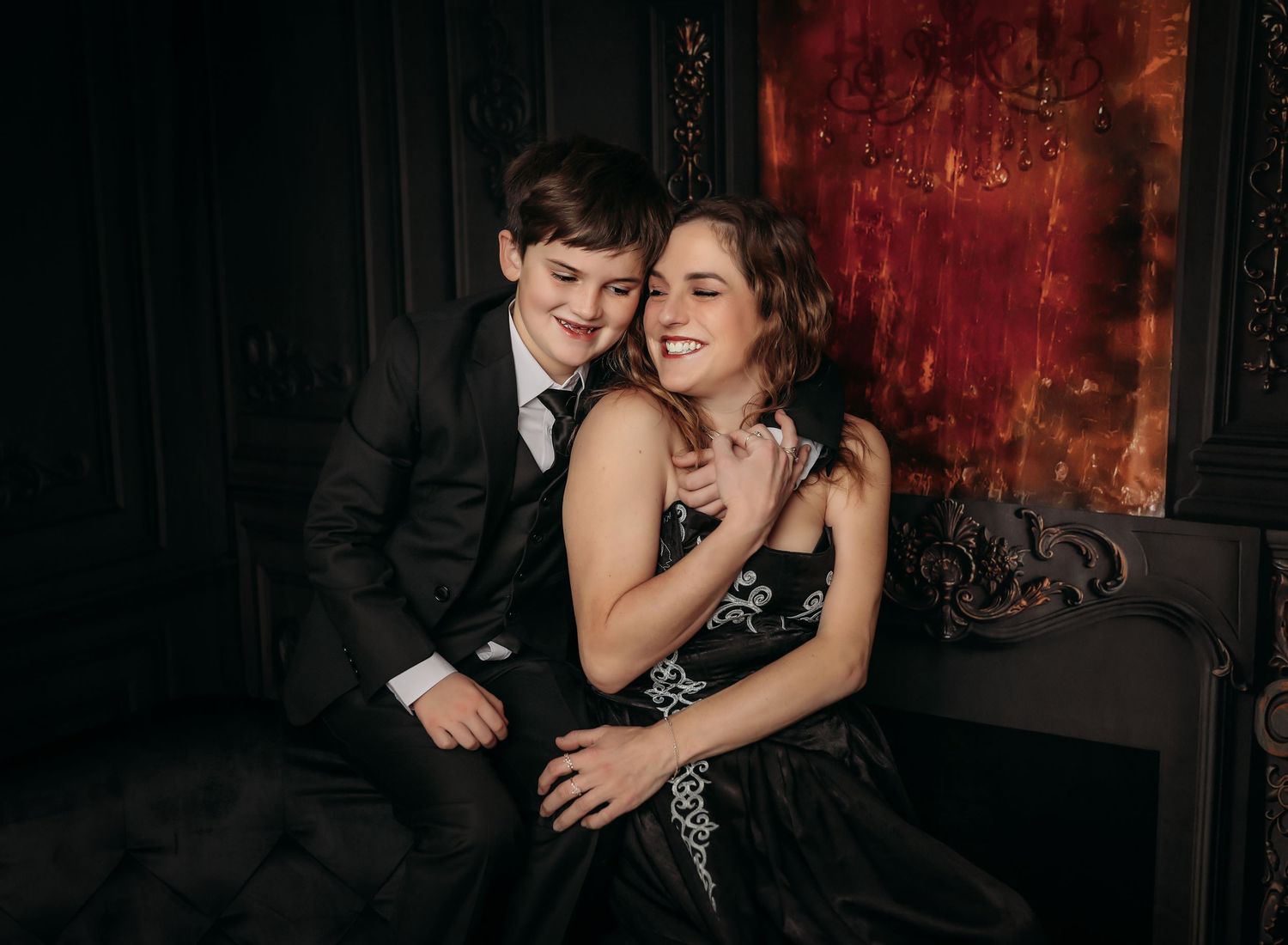 A romantic couple in formal attire embraces on a dark sofa against a red textured wall during an intimate photoshoot.