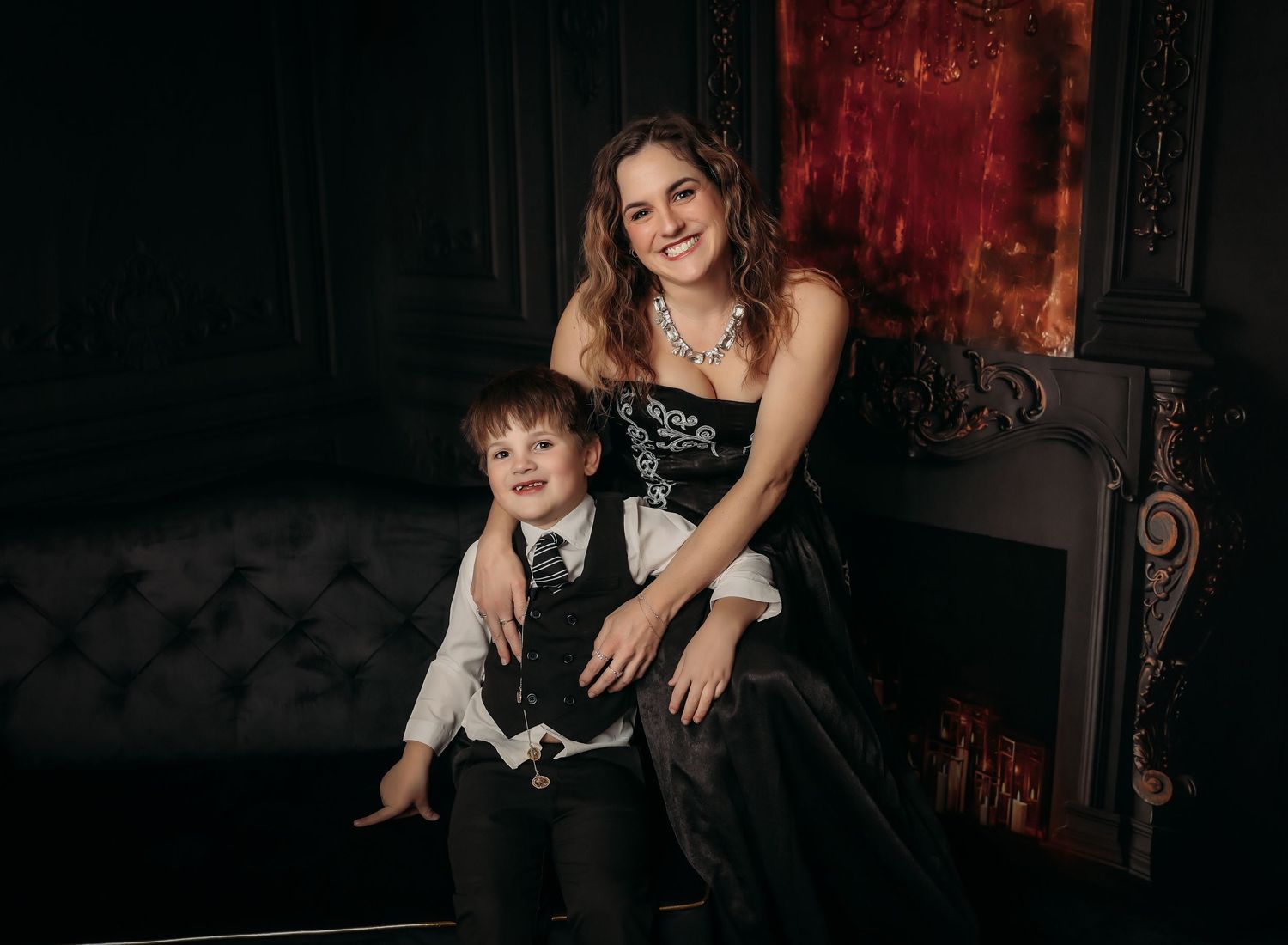 A mother and young son pose together on a dark tufted couch against a dramatic red and black backdrop.