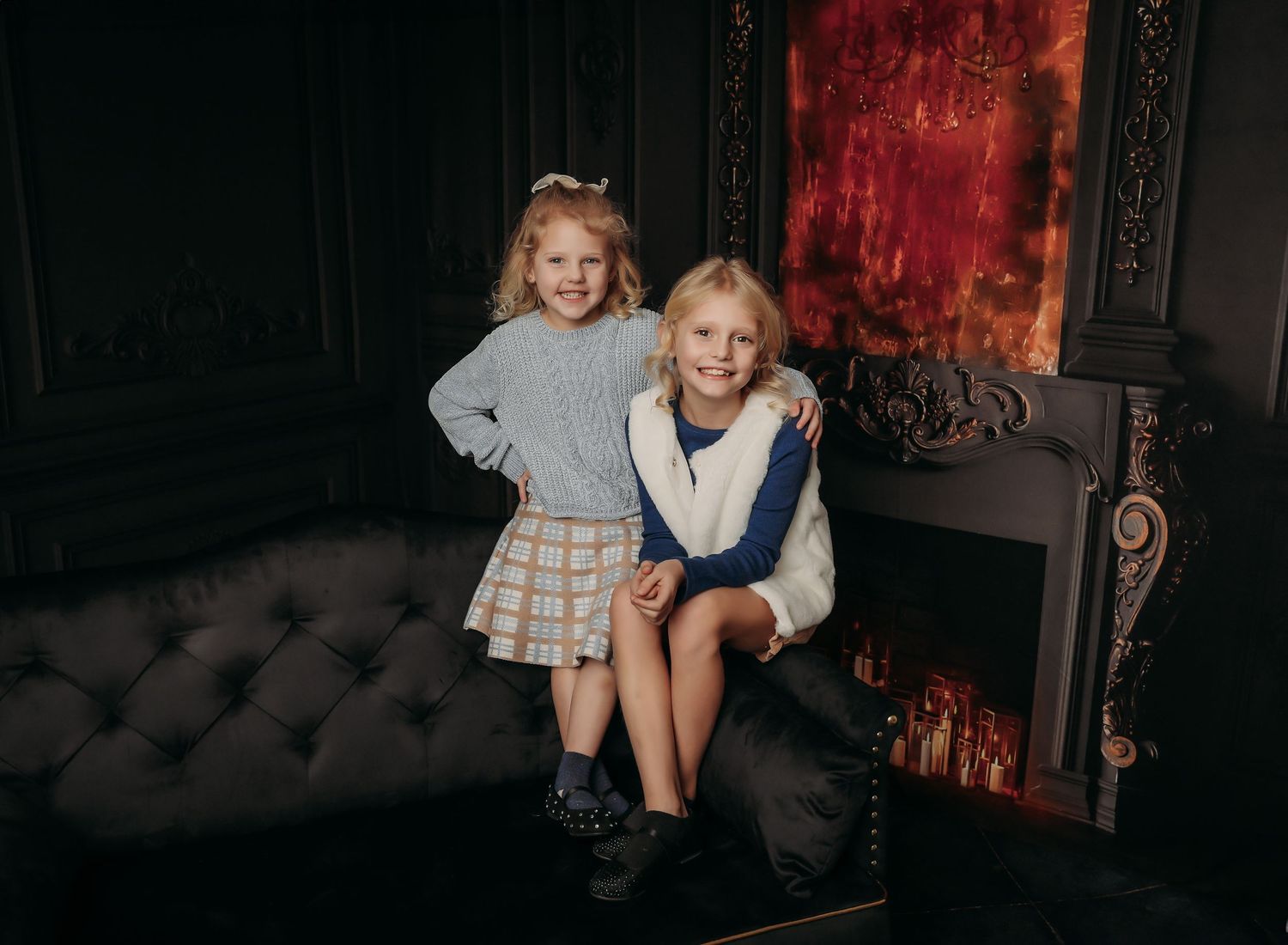 Two children sit together on a dark tufted sofa against a moody red backdrop in a vintage-style setting.