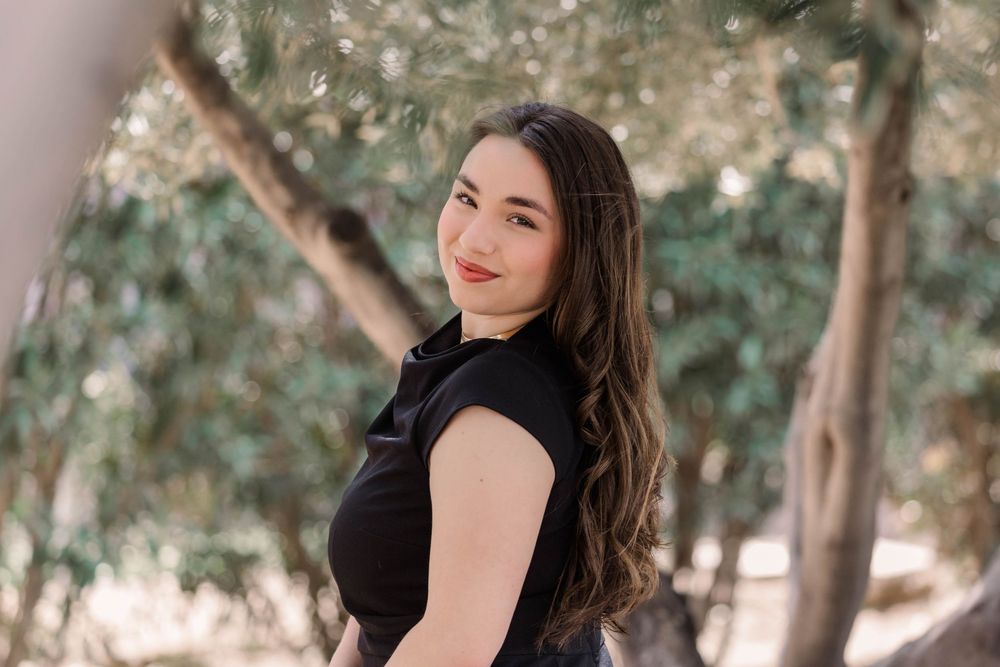 A person in a black dress poses outdoors against a natural background with trees and greenery.