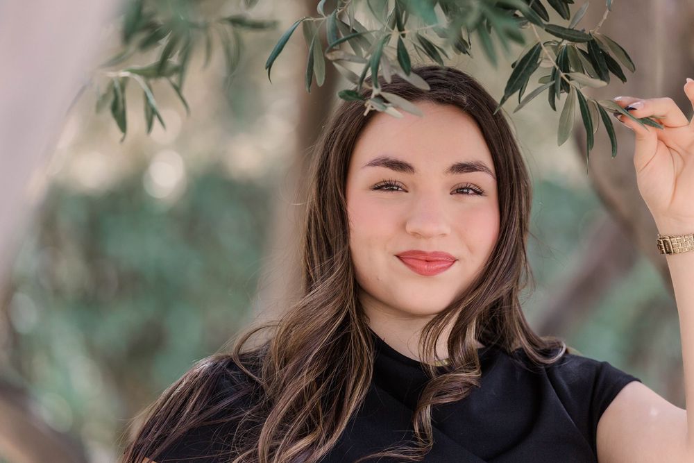 A person with long brown hair smiles outdoors under olive tree branches in natural lighting.