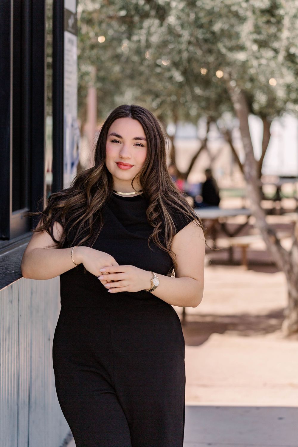 A lifestyle portrait of someone in an elegant black outfit standing against a wall with trees in background.