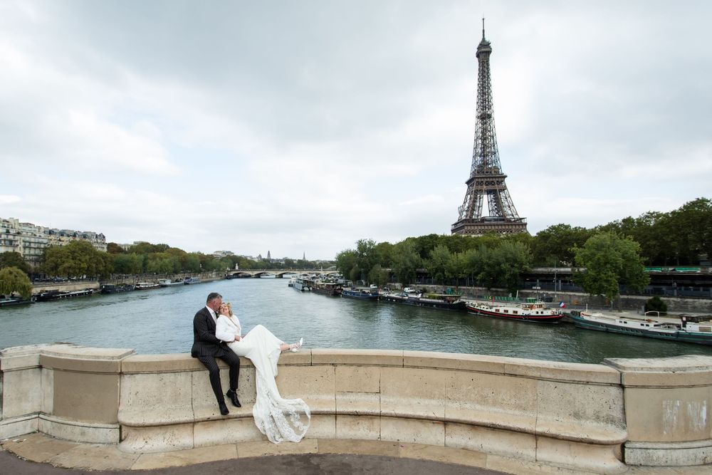 A couple in wedding attire sits on a stone bridge overlooking the Seine River with the Eiffel Tower in Paris.